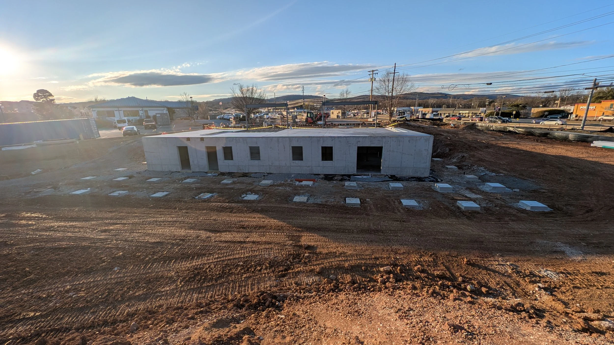 Construction site with a partially built concrete structure and numerous concrete foundation pads on the ground, with a parking lot and buildings in the background under a setting sun.