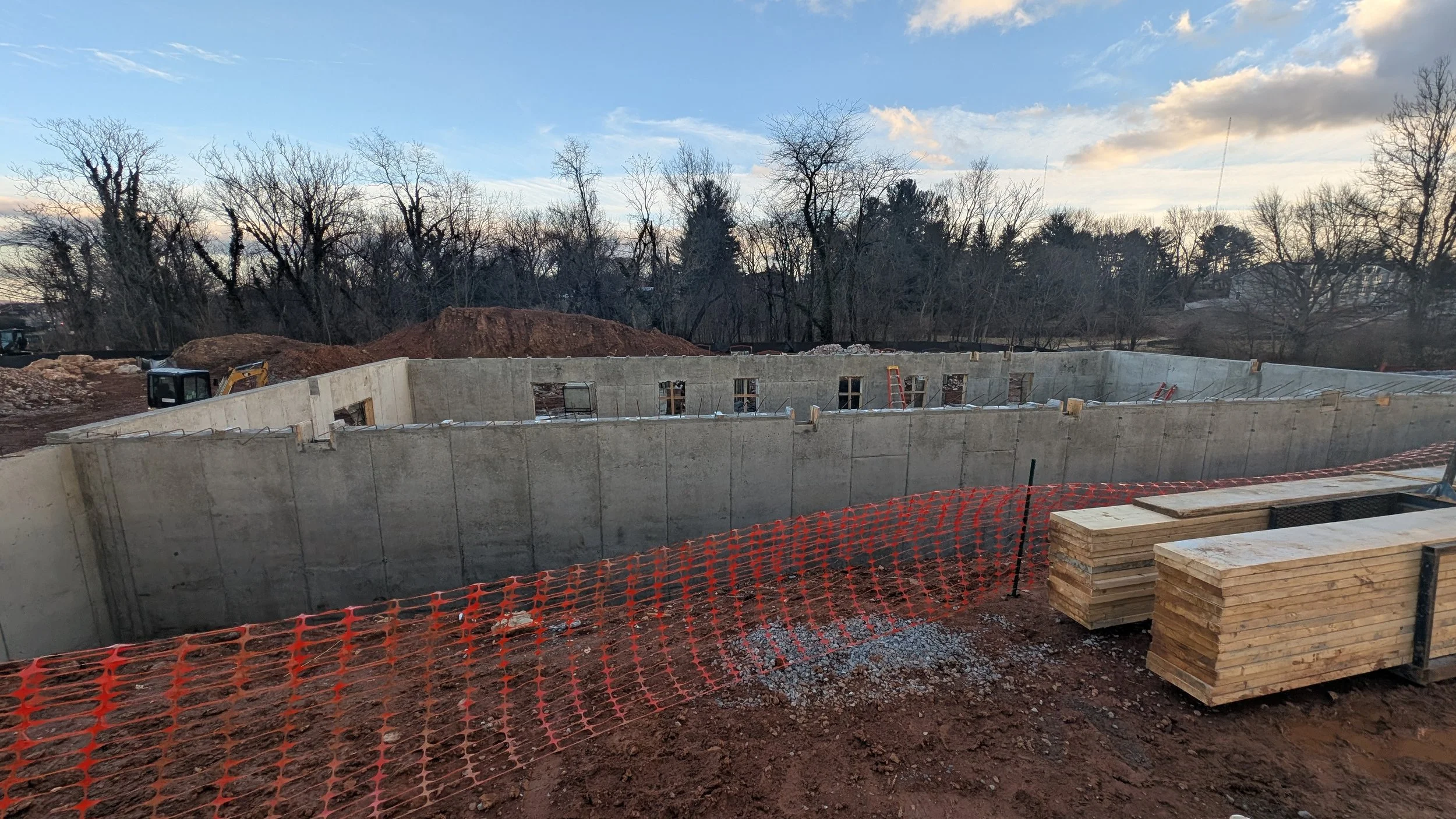 Construction site with concrete foundation walls, wooden planks, and orange safety fencing, with trees and a cloudy sky in the background.