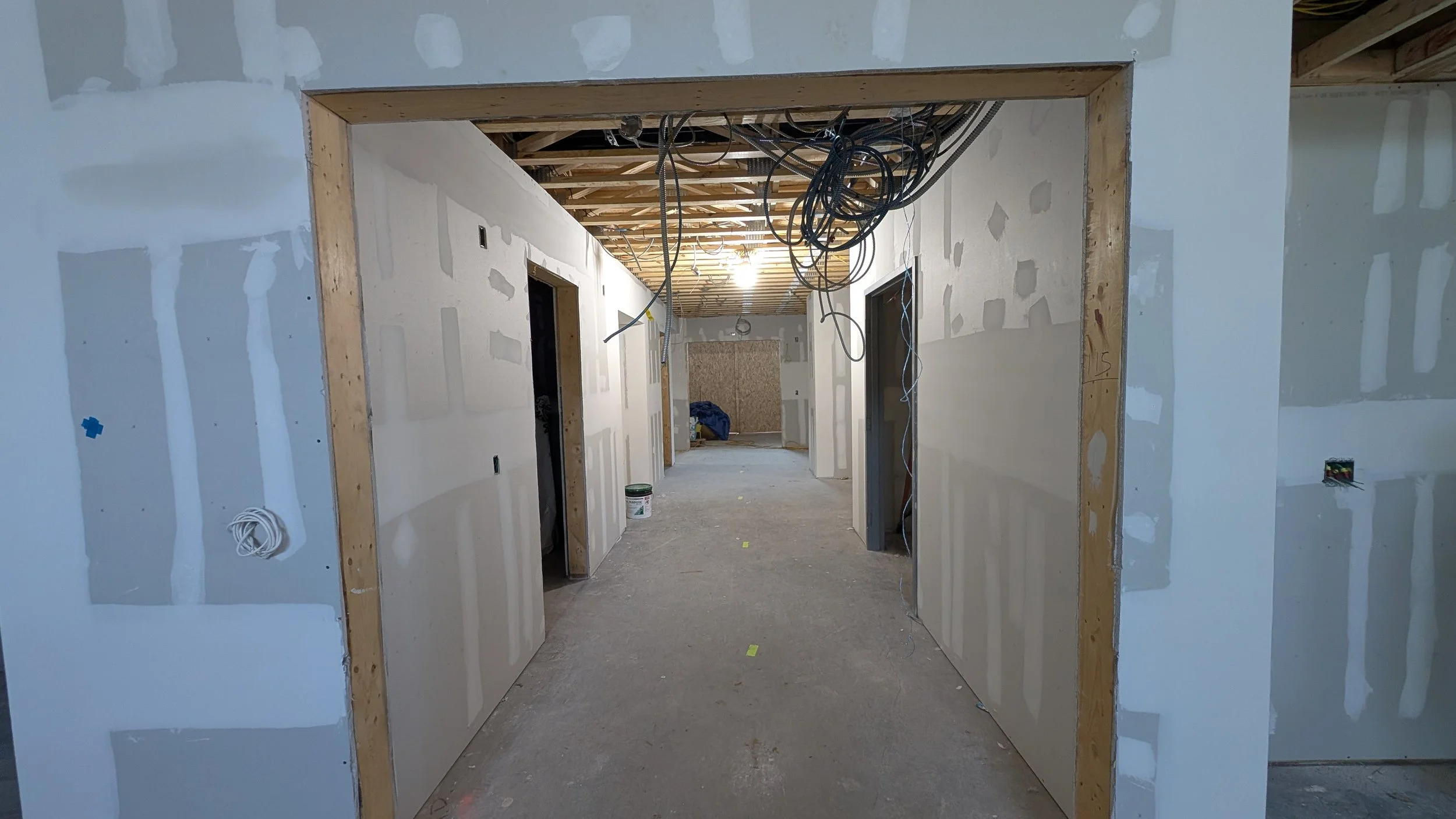 Interior of a building under construction with unfinished drywall walls and exposed ceiling with hanging electrical wires.