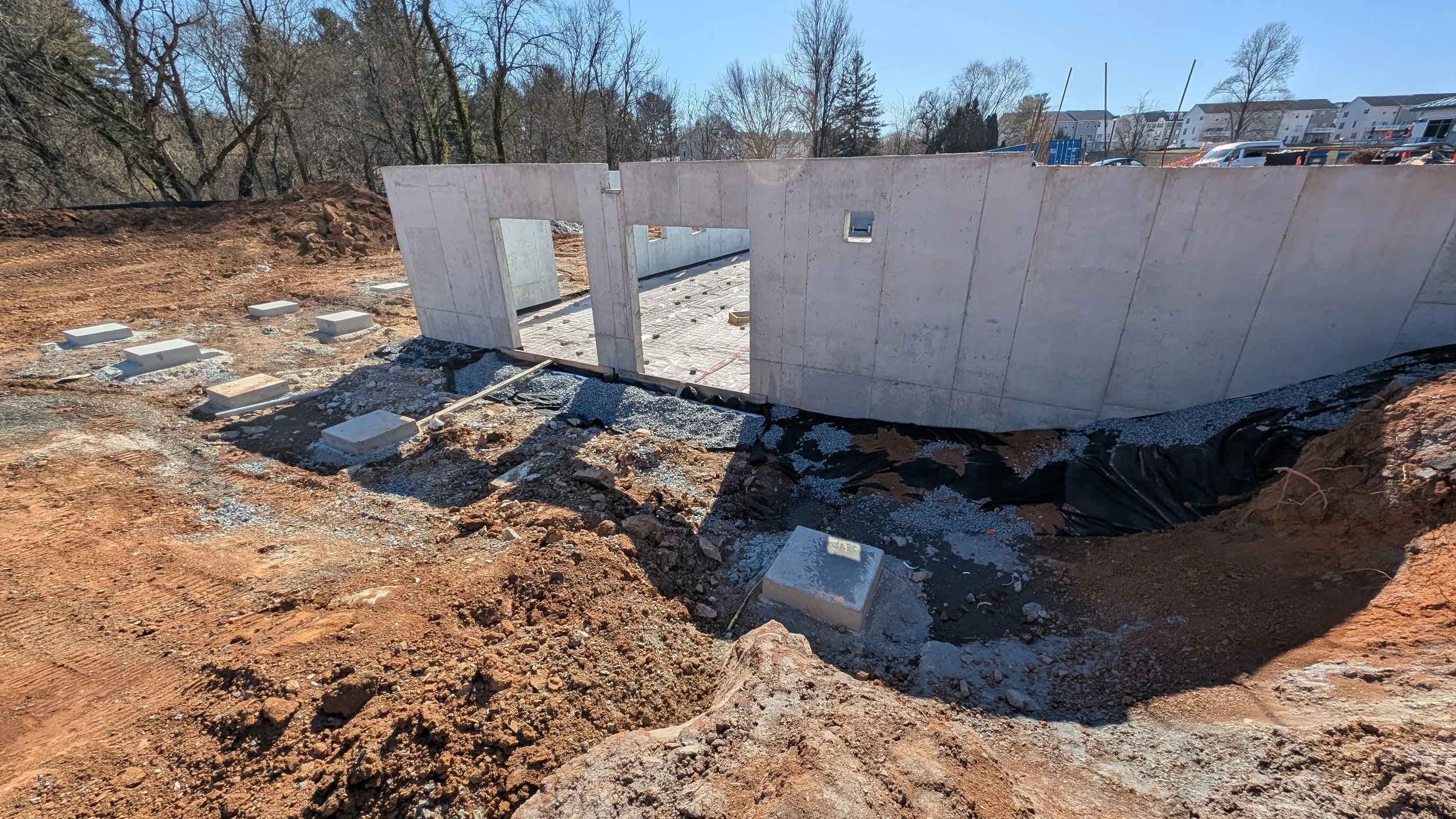 Construction site with a concrete wall, small windows, and a partially excavated area with gravel and dirt, in a residential area with trees and buildings in the background.
