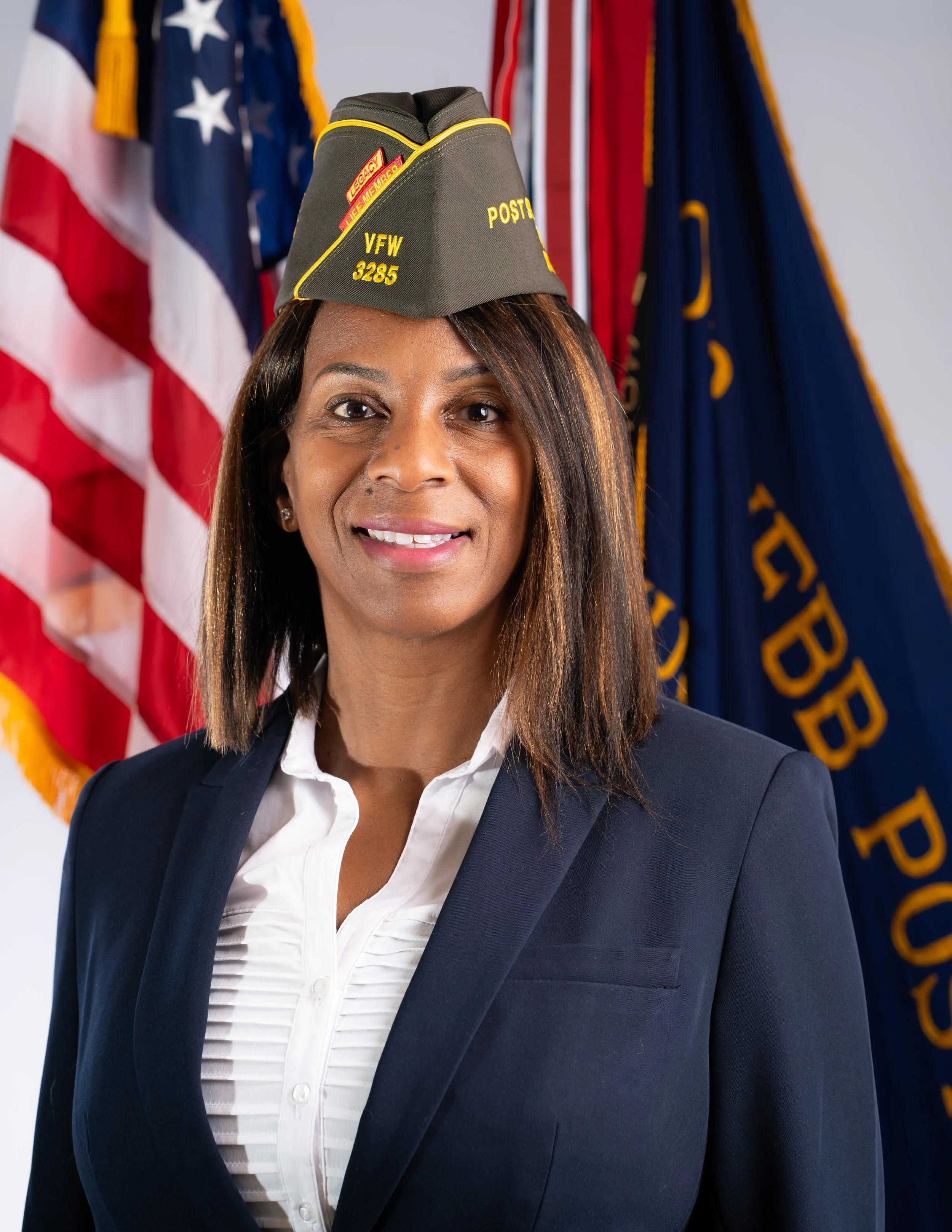 A woman with black hair wearing a VFW cap, black blazer, and earrings, standing next to an American flag, smiling at the camera.