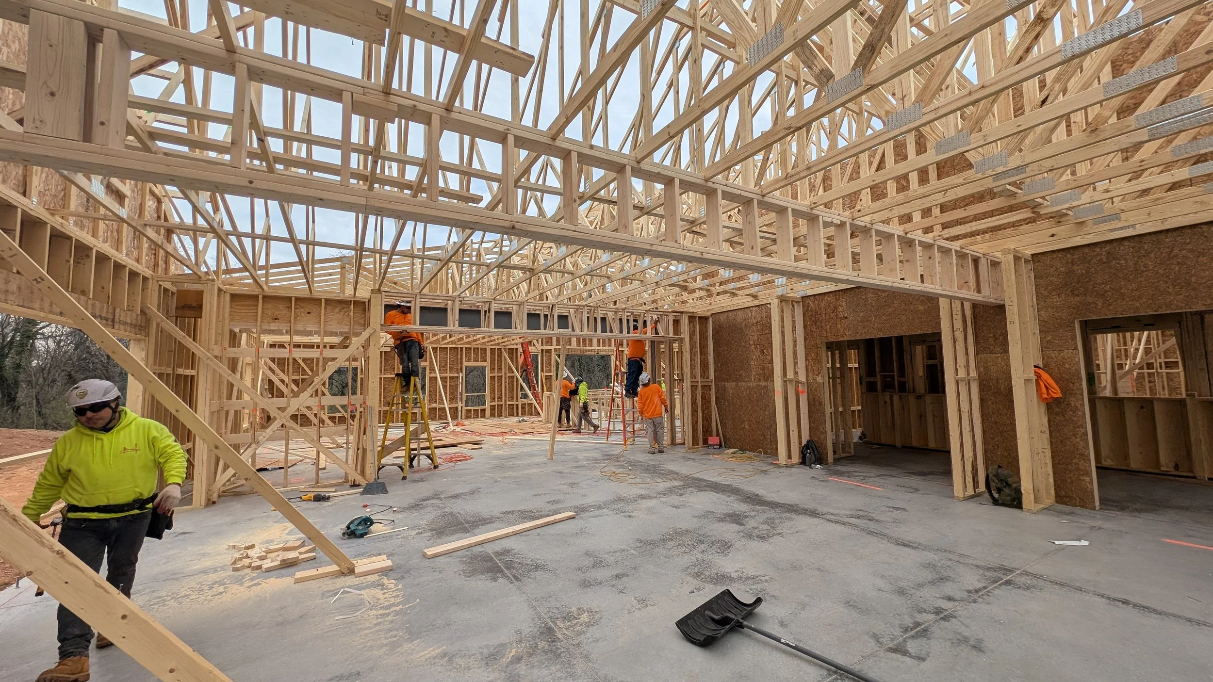 Construction workers building a wooden framework for a house, with exposed roof trusses and partial walls, on a cloudy day.