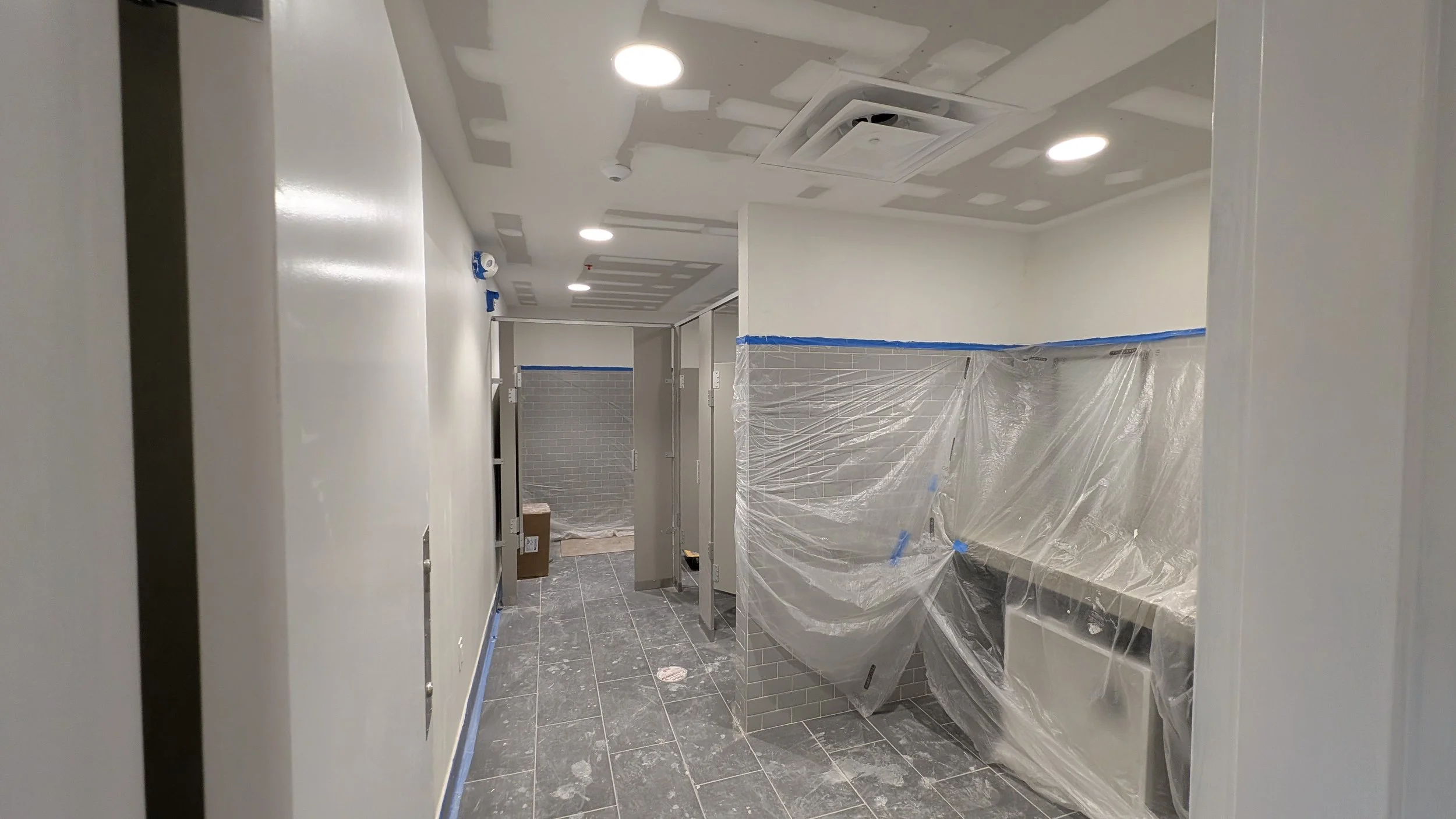Inside a commercial restroom under construction with plastic-covered sinks and partitions, and unfinished ceiling with recessed lighting.