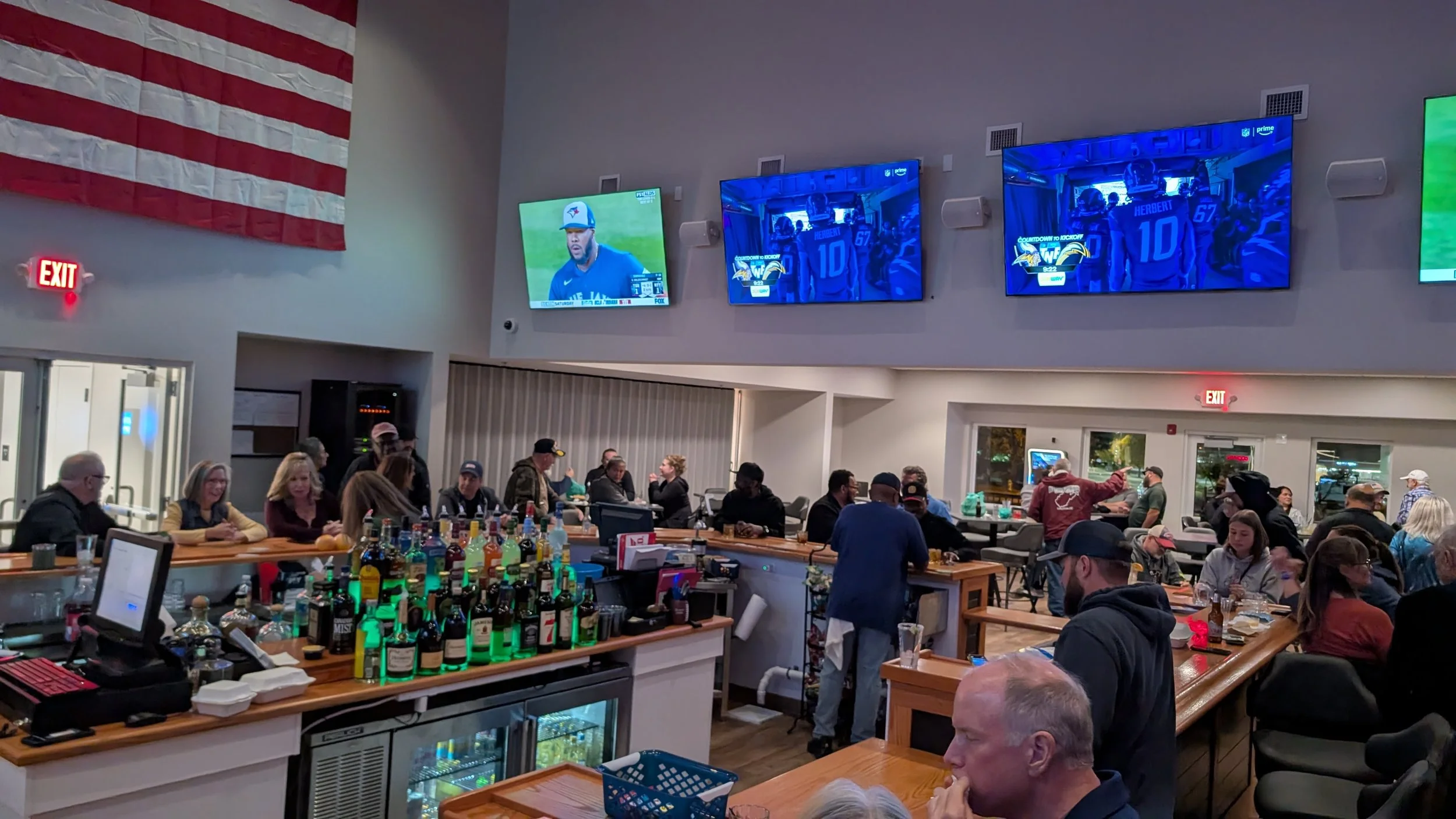 People watching sporting events on television in a bar with a basketball game on the screens, bar counter with bottles of alcohol, and American flag on the wall.