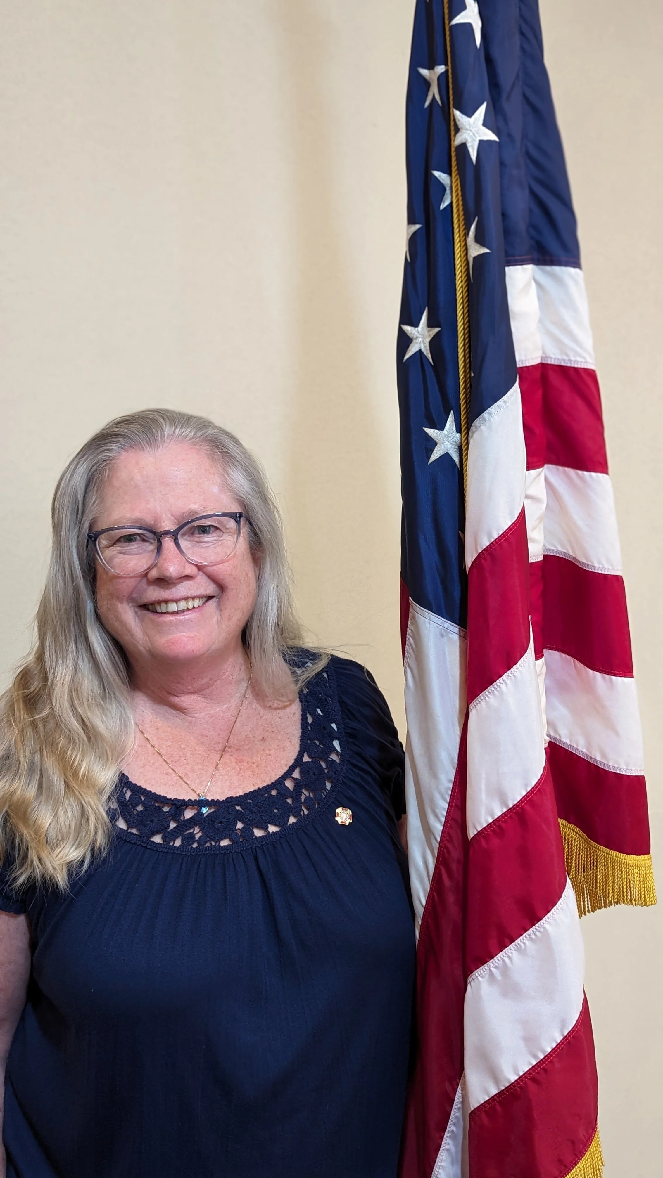 A smiling woman with glasses and long blonde hair standing next to a United States flag.