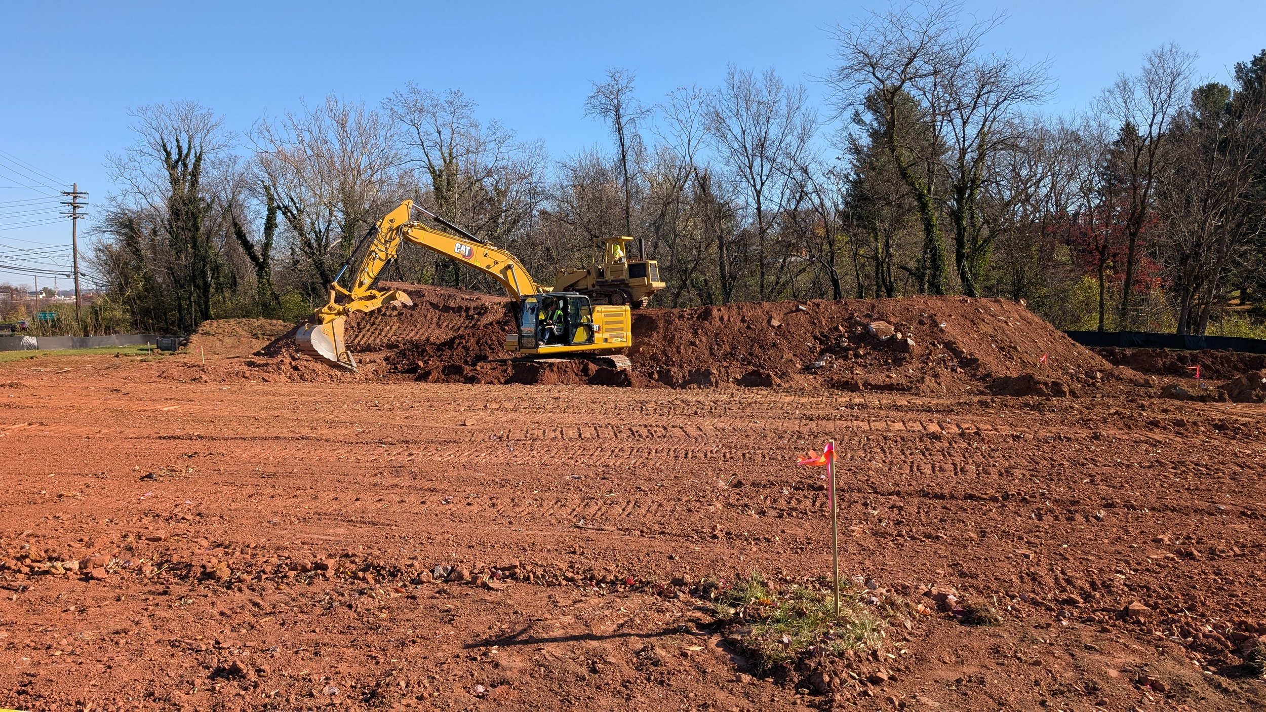 Construction site with bulldozer and excavator working on dirt mound, leafless trees in background, blue sky, and red flags marking the area.