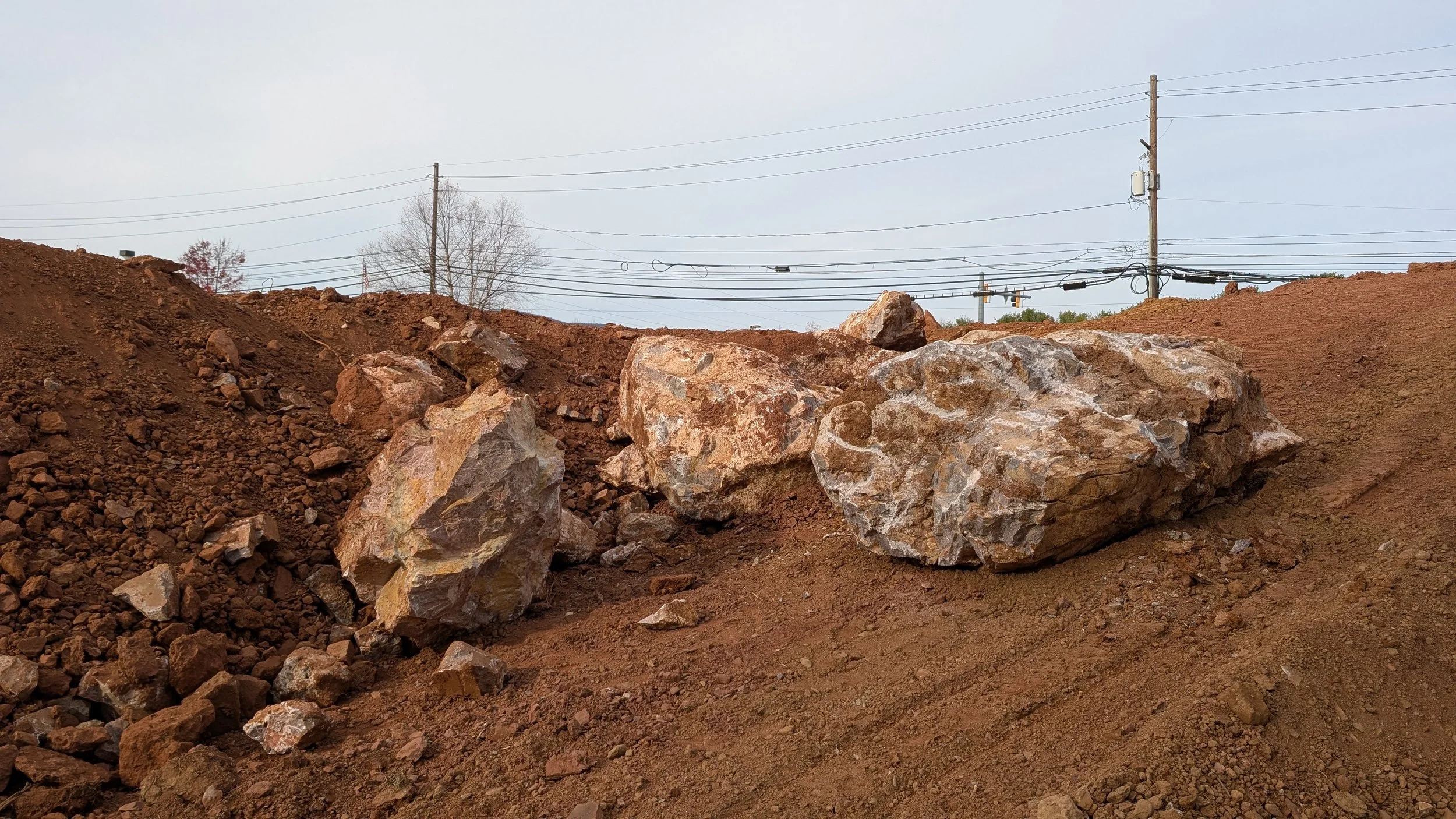 Large rocks on a disturbed dirt surface, with power lines and poles in the background under an overcast sky.