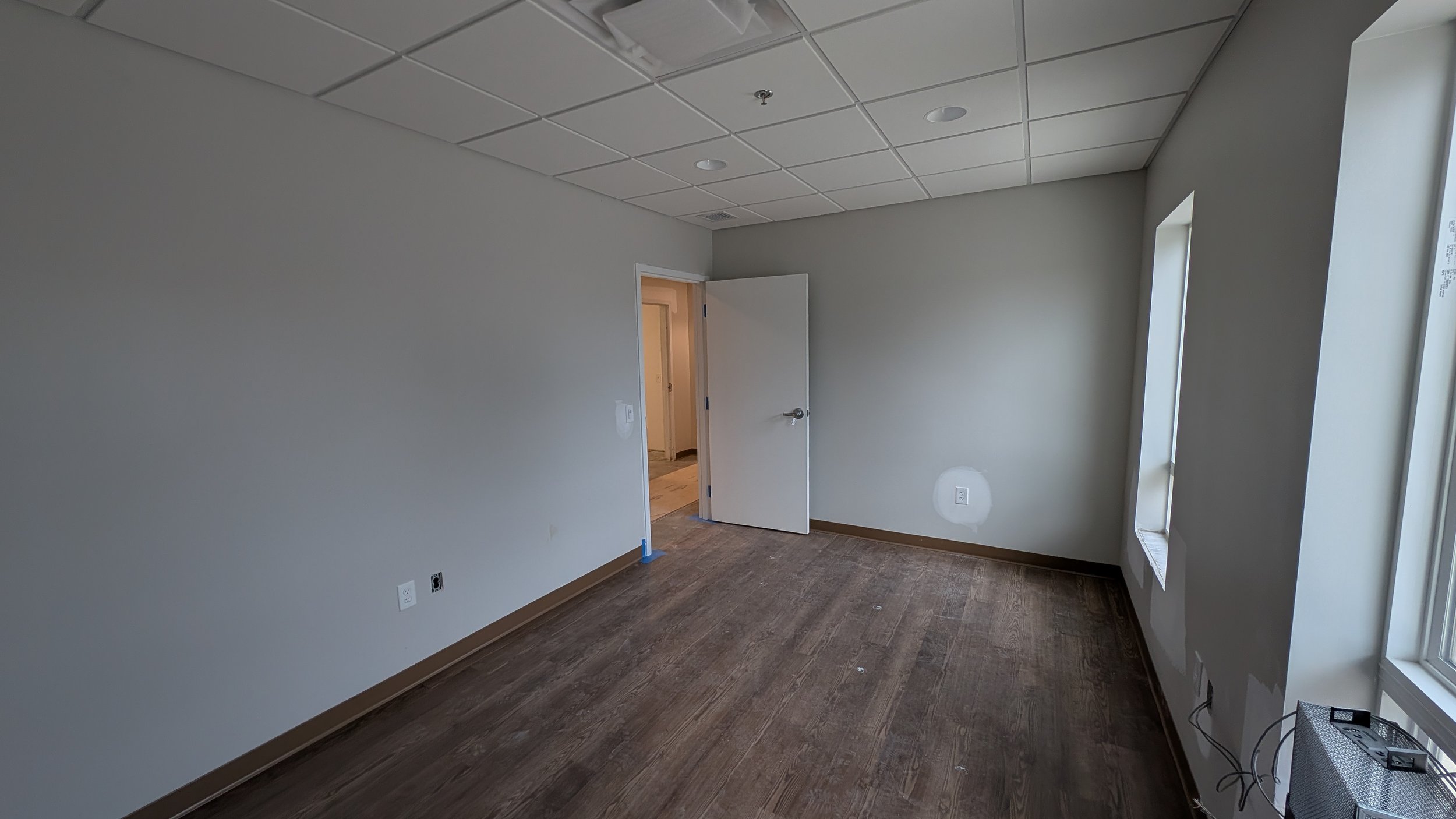 An empty room with beige walls, dark hardwood flooring, a white ceiling with recessed lighting, a window on the right side, and an open door leading to another room.