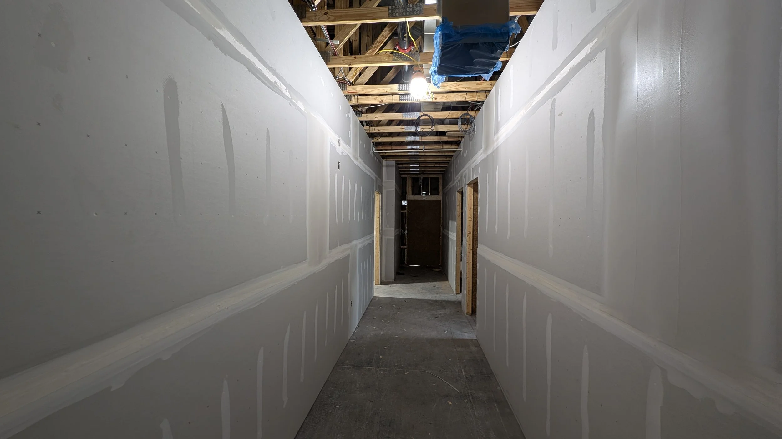 Construction hallway with drywall partially installed, exposed ceiling with wooden beams and electrical wiring, and a construction light illuminating the area.