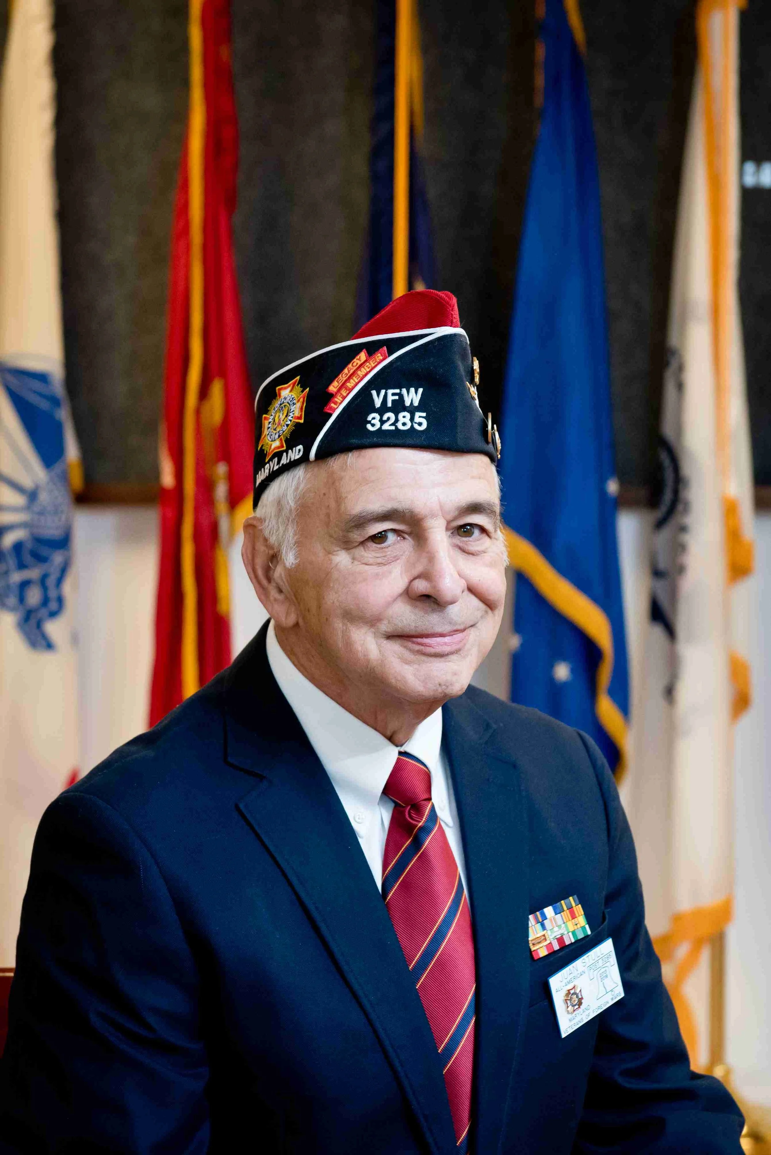 An elderly man in a navy blazer and a red striped tie, wearing a black veterans hat, standing in front of a backdrop of flags.