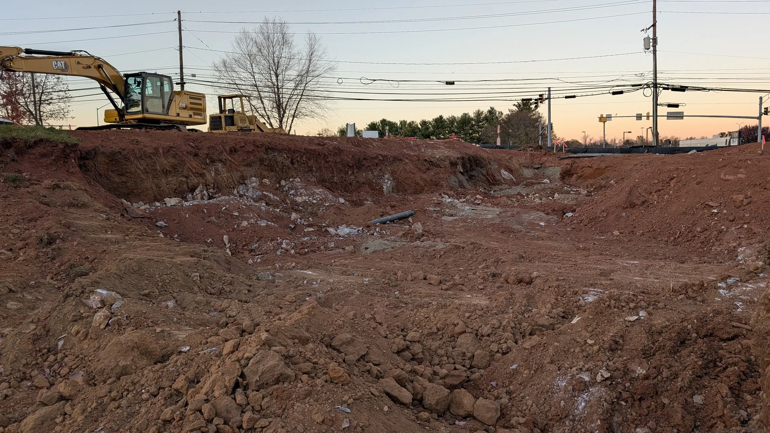 Construction site with excavator and deep dirt pit at sunset, with power lines and trees in the background.