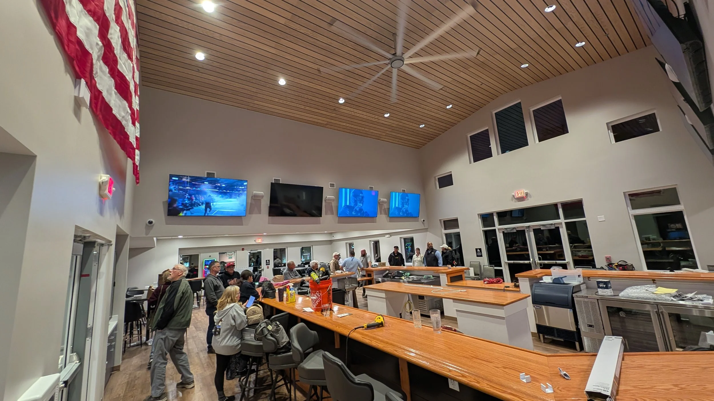 People inside a modern lobby or waiting area with TV screens on the wall, a wooden ceiling, and an American flag hanging on the wall.