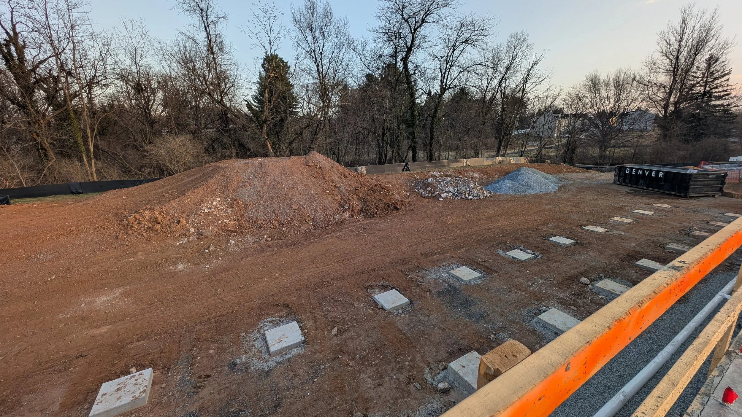 A construction site with dirt mounds, gravel, and concrete slabs, with trees and houses in the background, and orange safety barriers in the foreground.