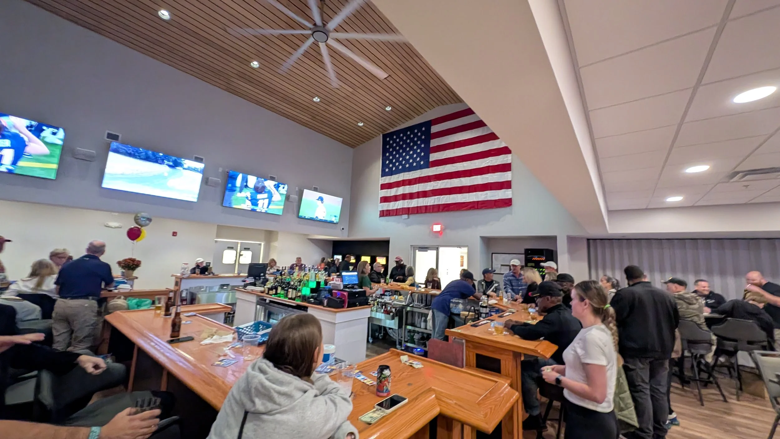 A lively bar and lounge area with people socializing and watching sports on multiple flat-screen TVs. The American flag hangs prominently on the wall, with a ceiling fan and wooden ceiling accents.