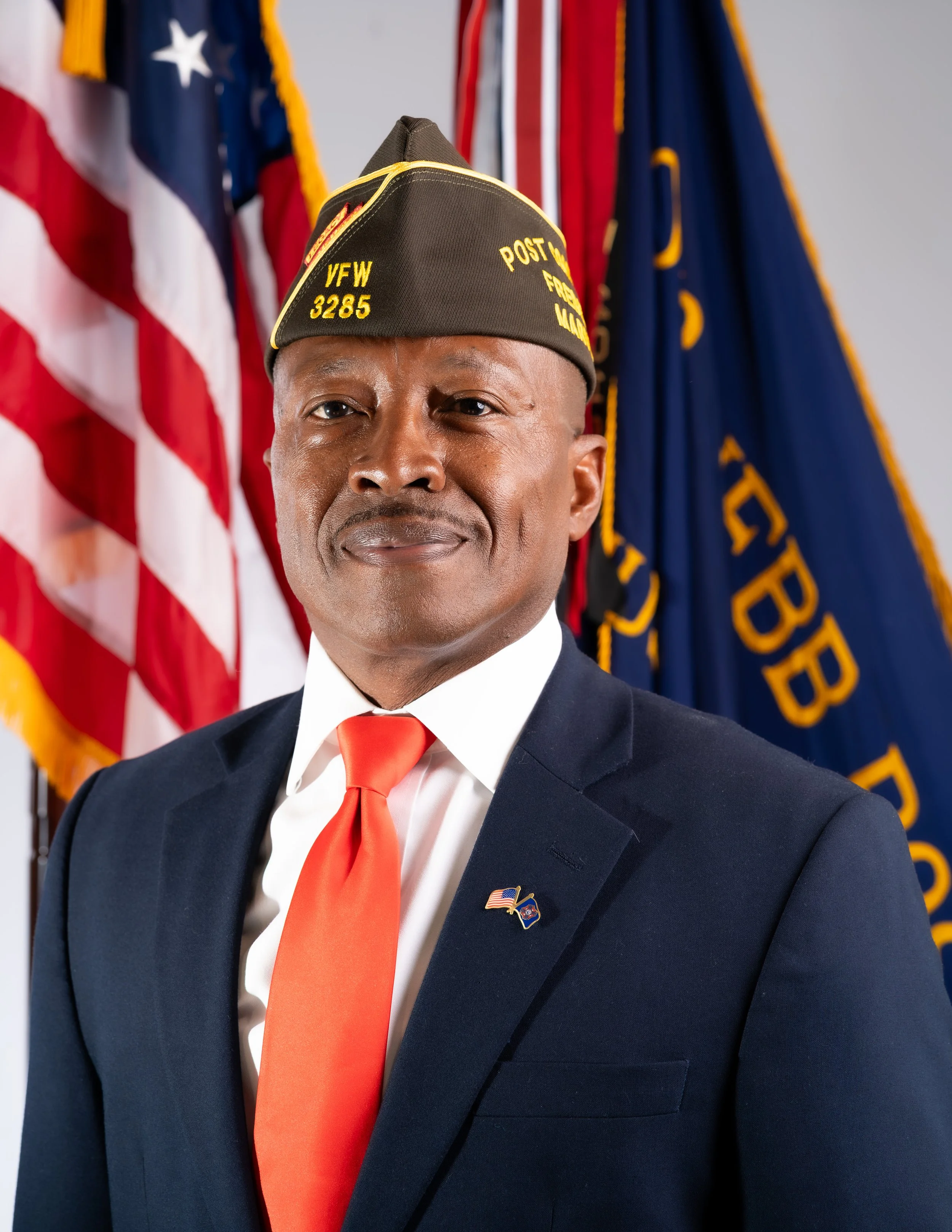A man wearing a black Veterans of Foreign Wars (VFW) cap with yellow lettering and a gray beard, smiling in front of an American flag.