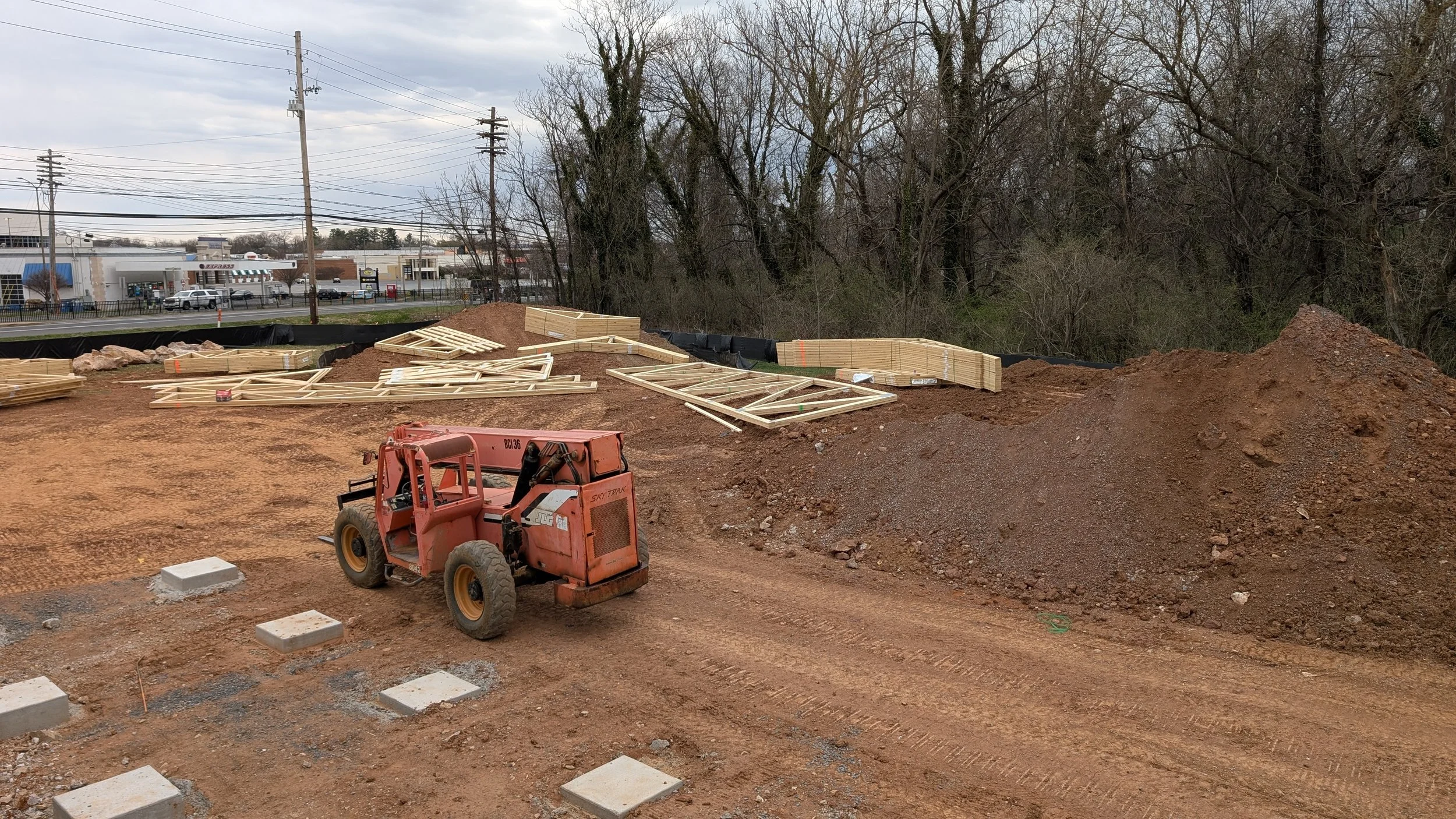 Construction site with wooden framing, a small red excavator, and piles of dirt, with trees and power lines in the background.