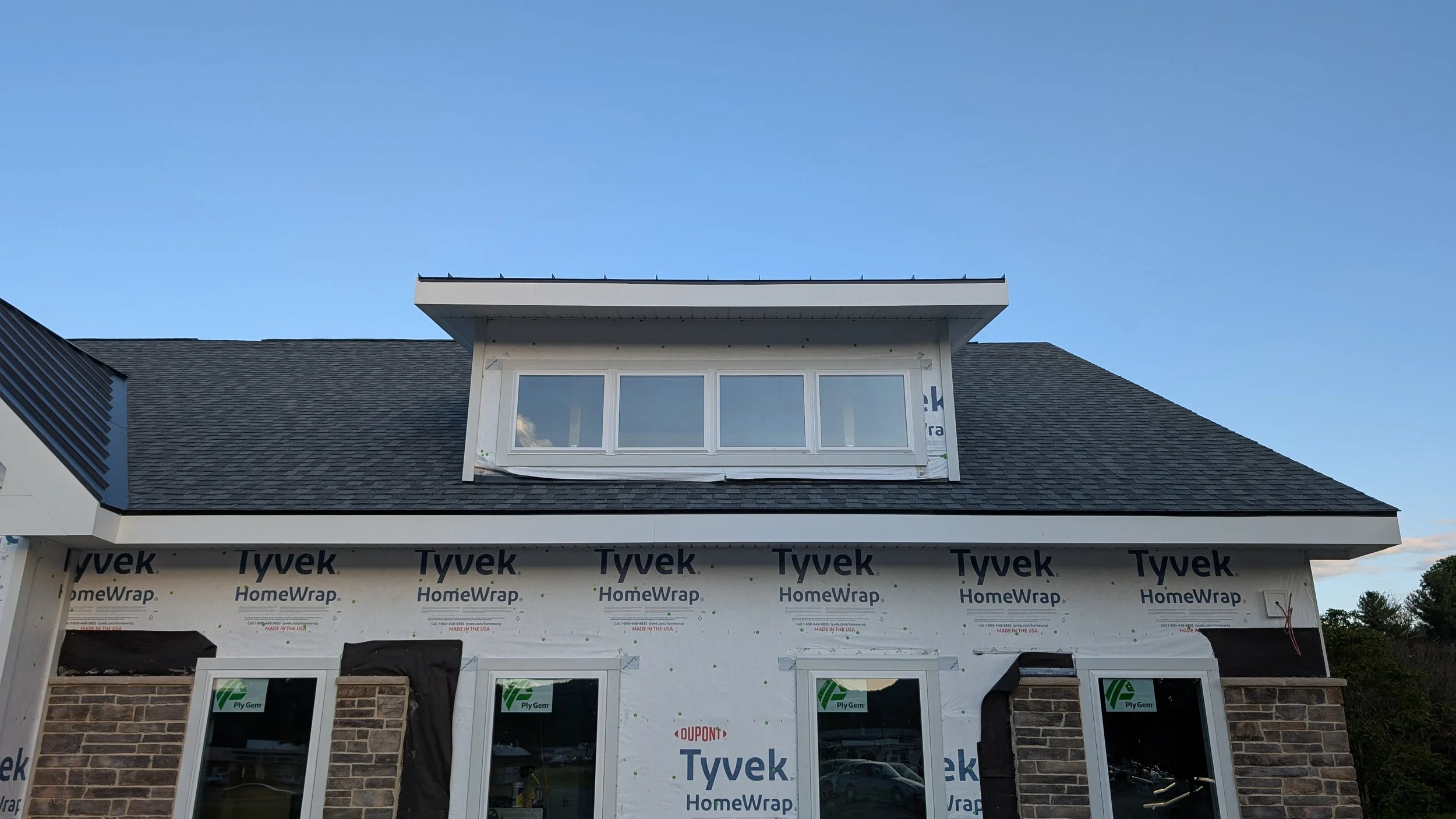 A house under construction with a partially finished exterior wall, featuring two large windows, brickwork, and a roof with a dormer window. The exterior is covered in Tyvek HomeWrap, and some building materials and wires are visible.