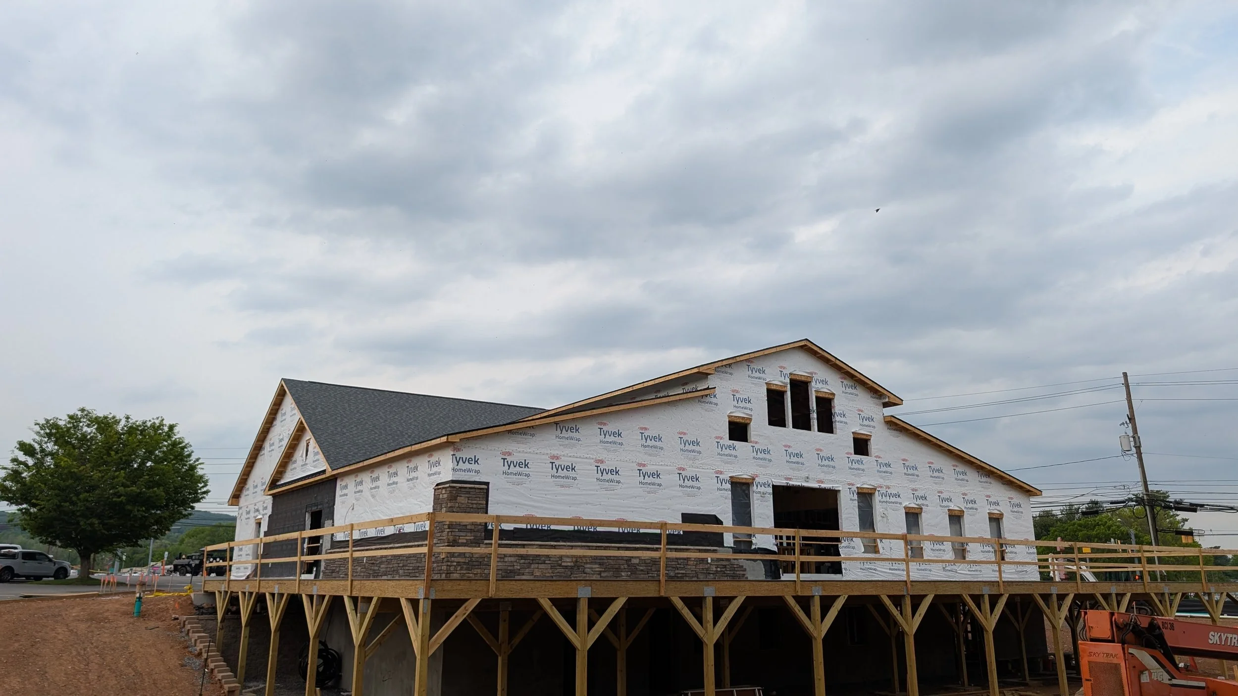 Under construction house with wooden frame and Tyvek house wrap, few windows and a dark shingled roof, on a raised foundation with support beams