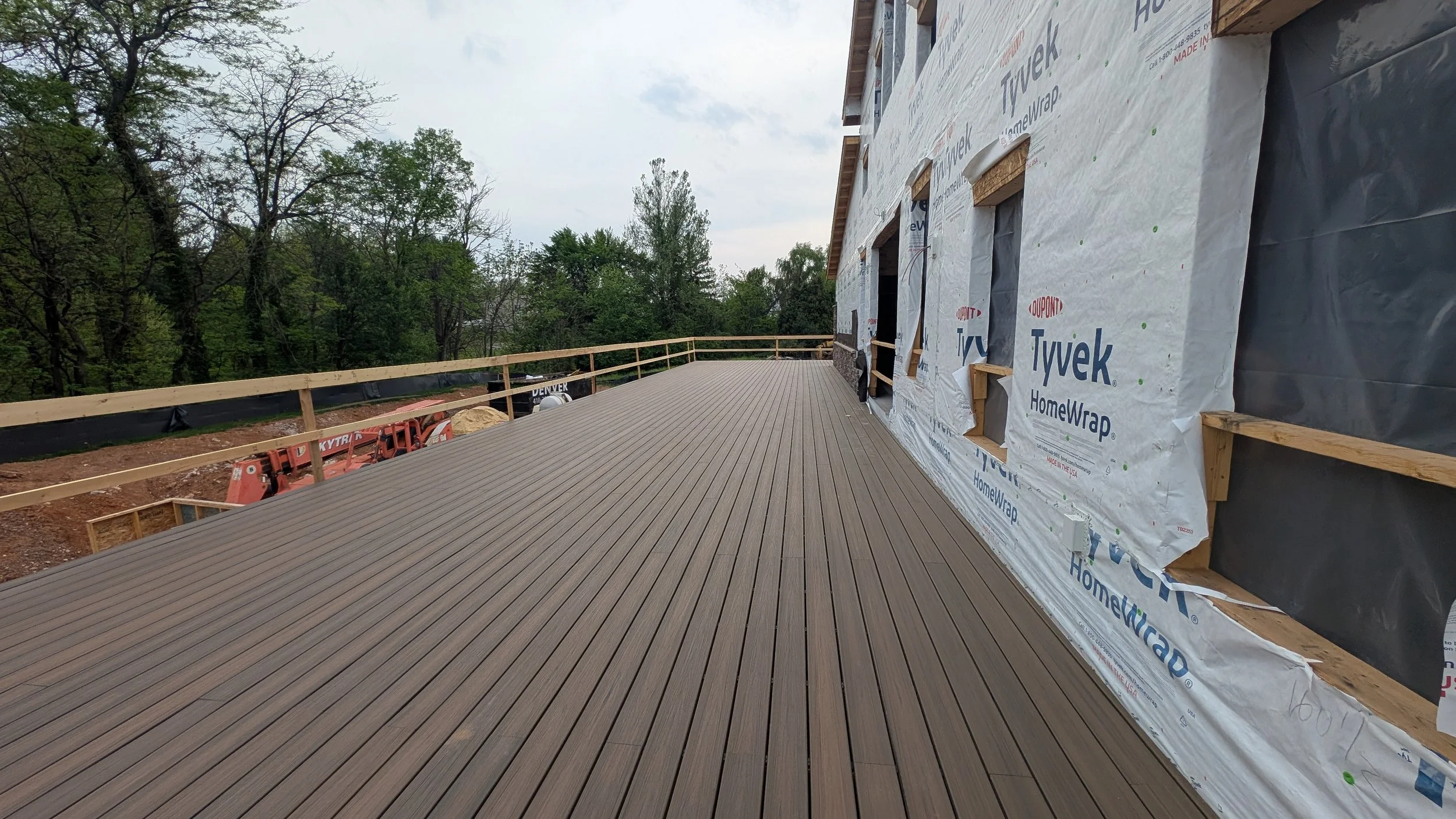 Construction site with a new wooden deck and a house under renovation covered in white Tyvek HomeWrap, surrounded by trees.