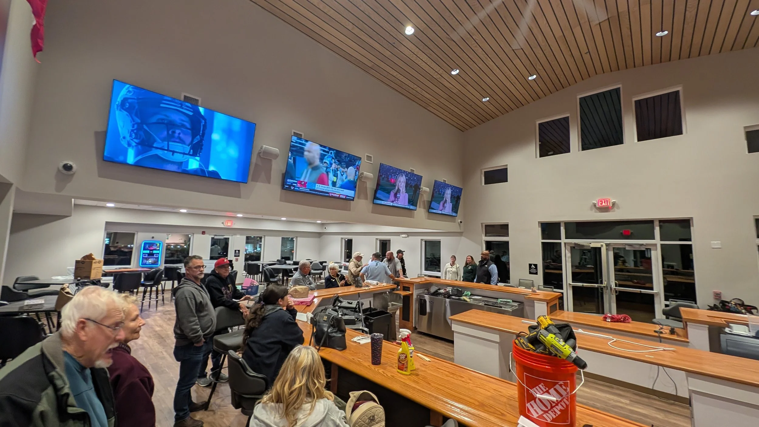 People gathering in a spacious room with multiple wall-mounted televisions showing sports and news, a wooden ceiling, and a bar area with tables and chairs.
