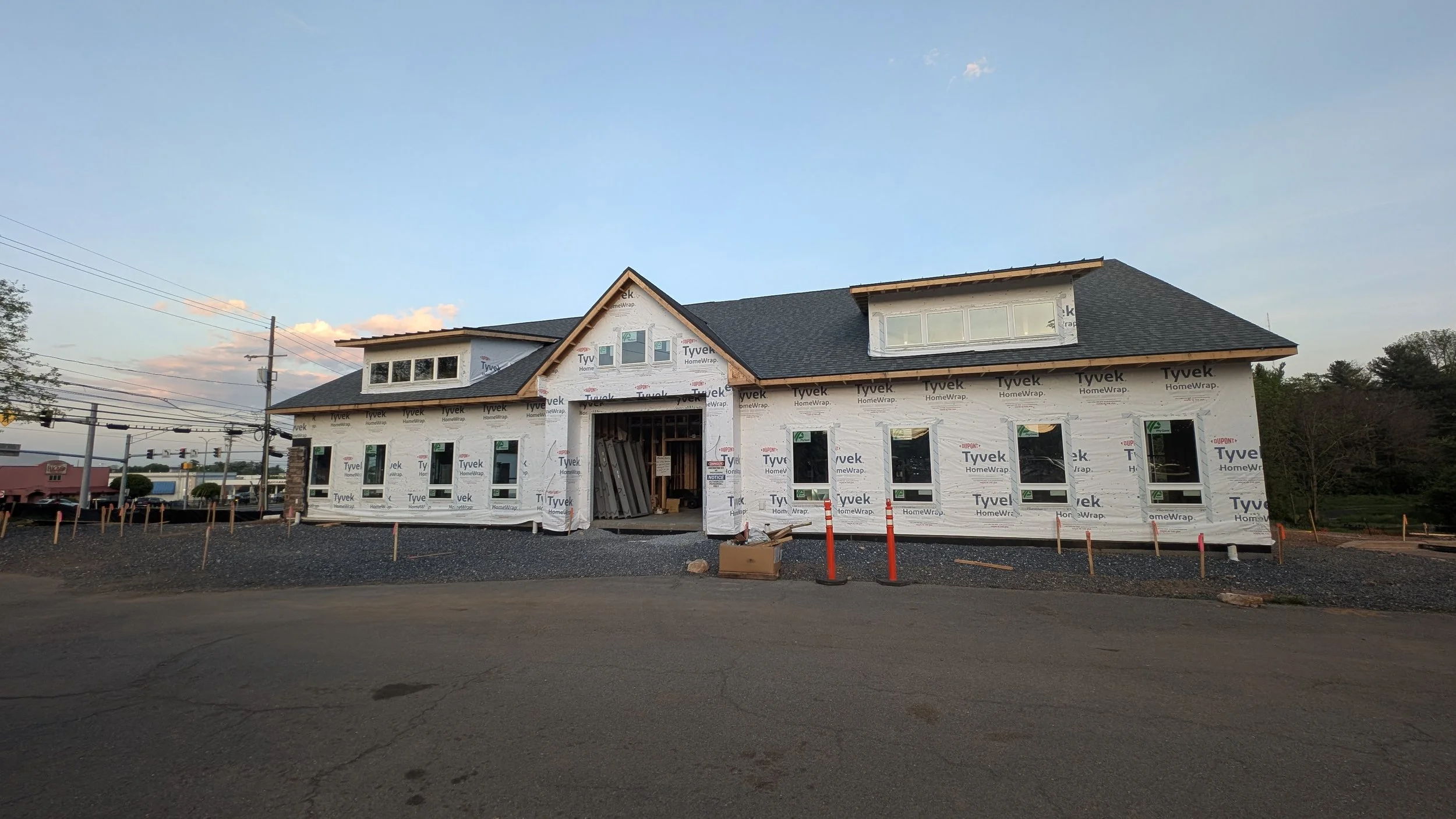 Under-construction building wrapped in white Tyvek house wrap, with several windows installed, a multi-gabled roof, and construction materials and orange safety posts in front.