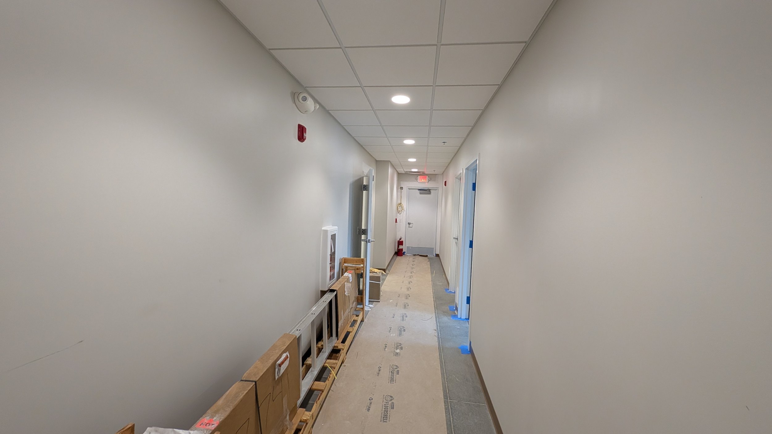 Empty hallway in a building under construction or renovation with boxes and tools on the floor, ceiling lights, and a fire extinguisher at the end.