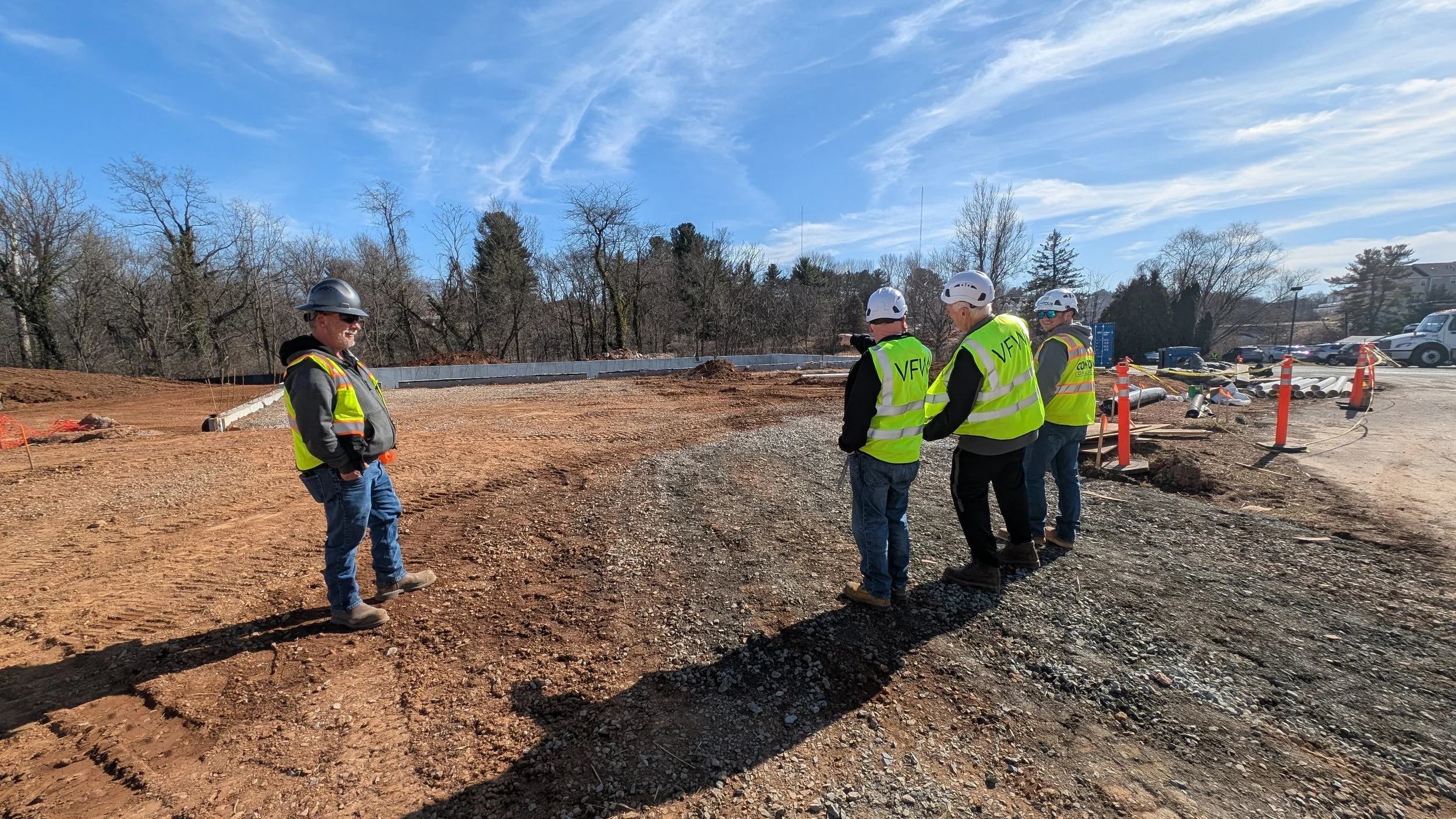 Construction workers wearing safety vests and helmets standing on a dirt site, discussing plans, with construction equipment and pipes visible nearby, and trees in the background under a blue sky.
