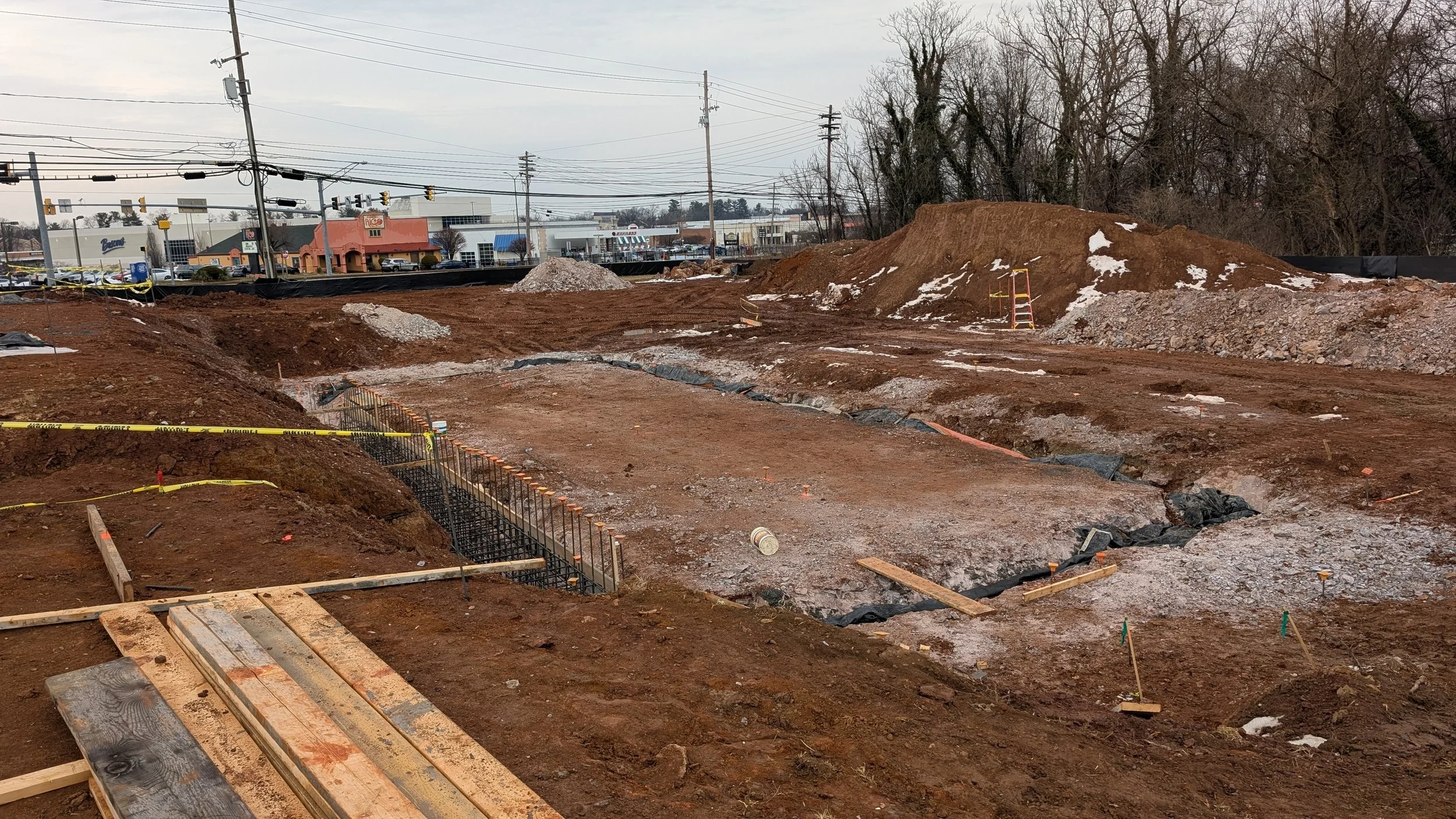 Construction site with exposed dirt, trenches, and building foundation prep, with nearby commercial buildings and power lines in the background.