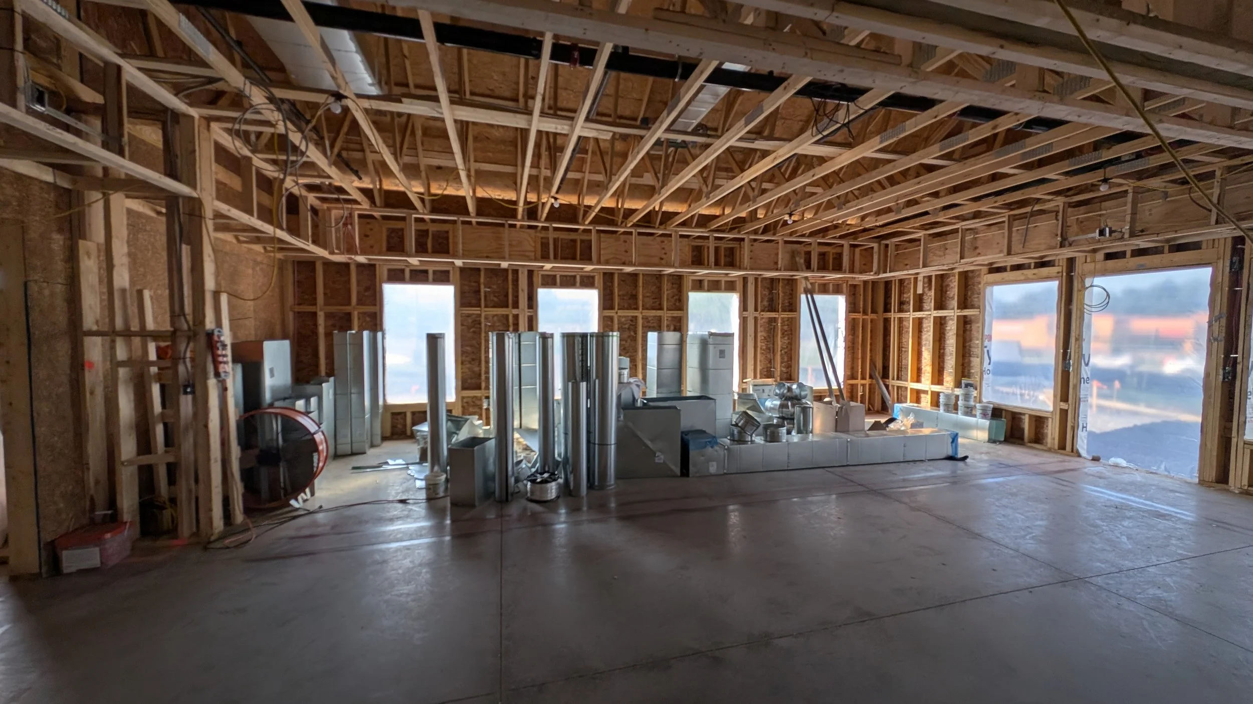 Interior view of a building under construction with exposed wooden framing, unfinished walls, and large windows. Construction materials and HVAC ductwork are visible on the floor.
