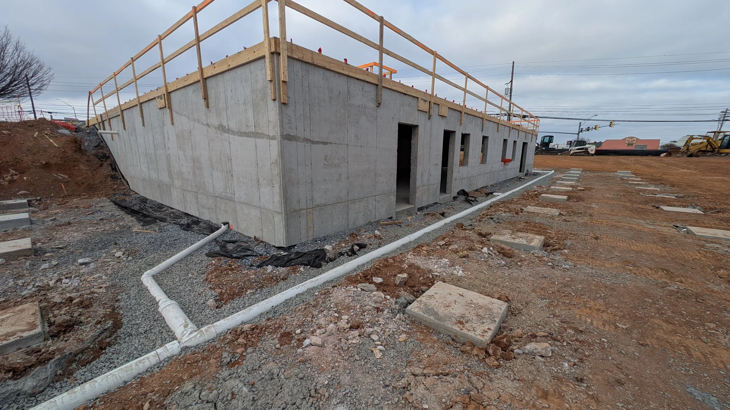 Construction site with a concrete building foundation, exterior walls, and open doorways; wooden railing on top and construction equipment in the background.