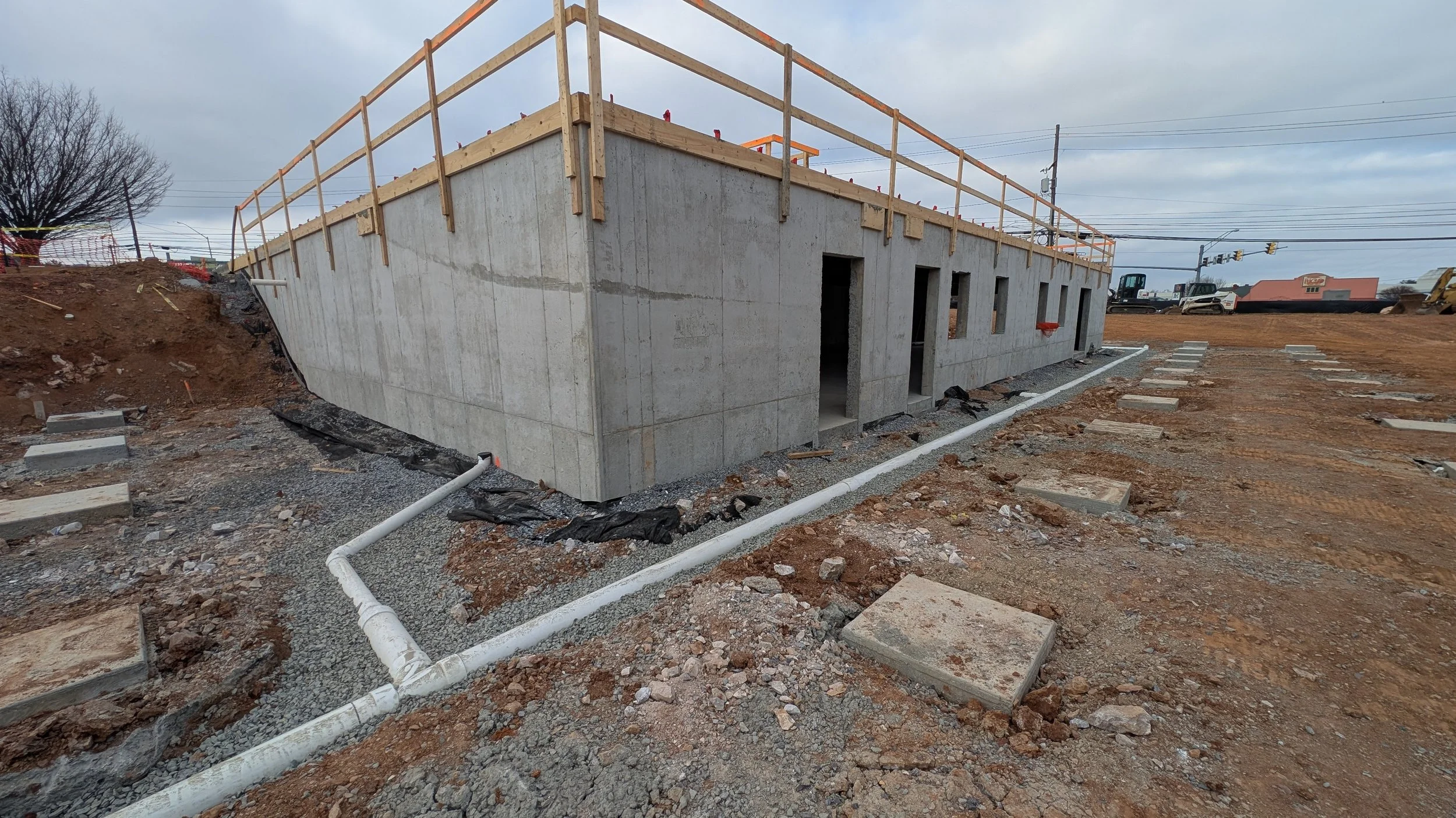 Construction site with a concrete building foundation and wooden railing on top, dirt and gravel surrounding, some concrete slabs and pipes on ground