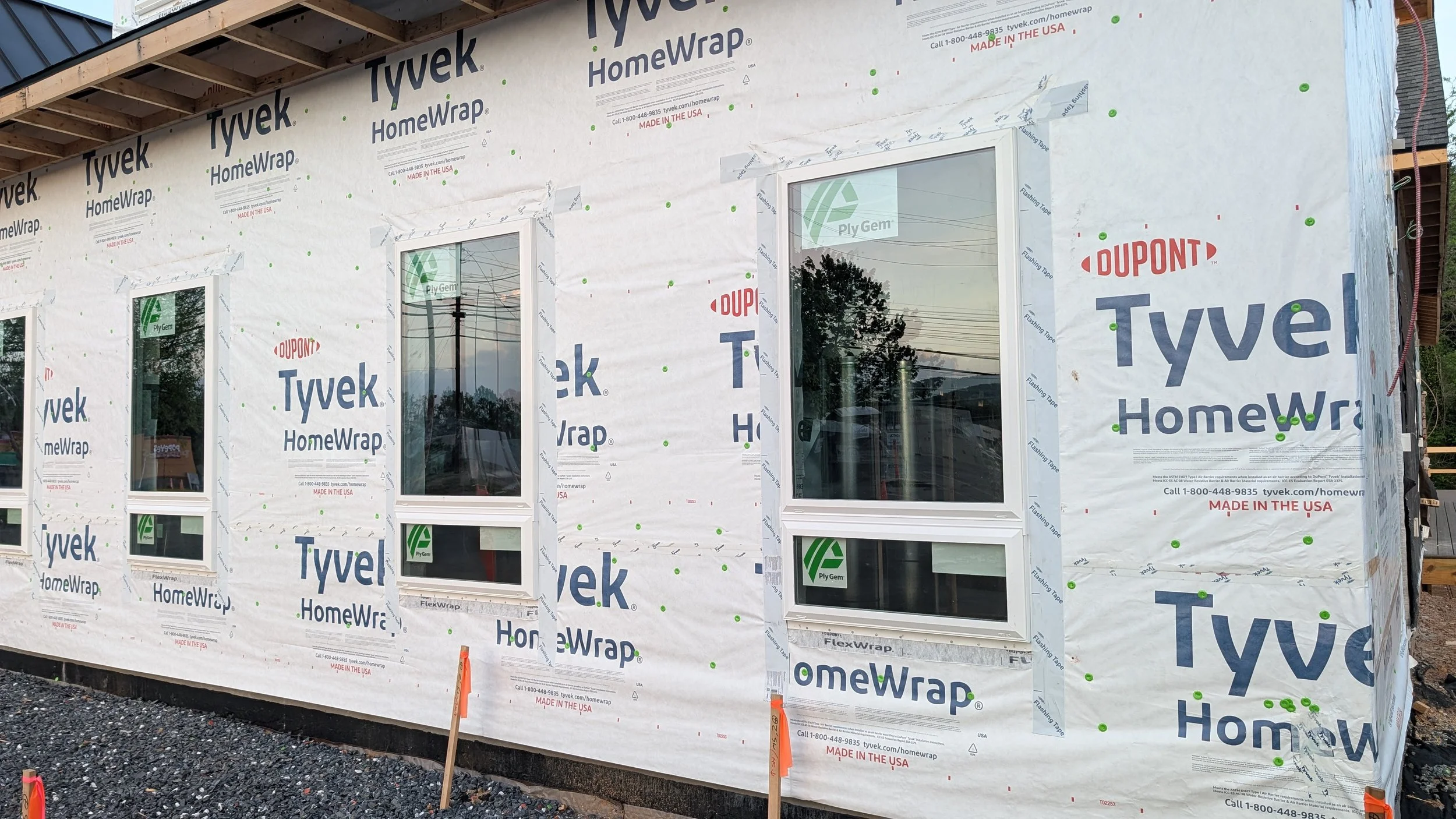 Construction site with a building wrapped in Tyvek HomeWrap material, three windows installed, and gravel on the ground.