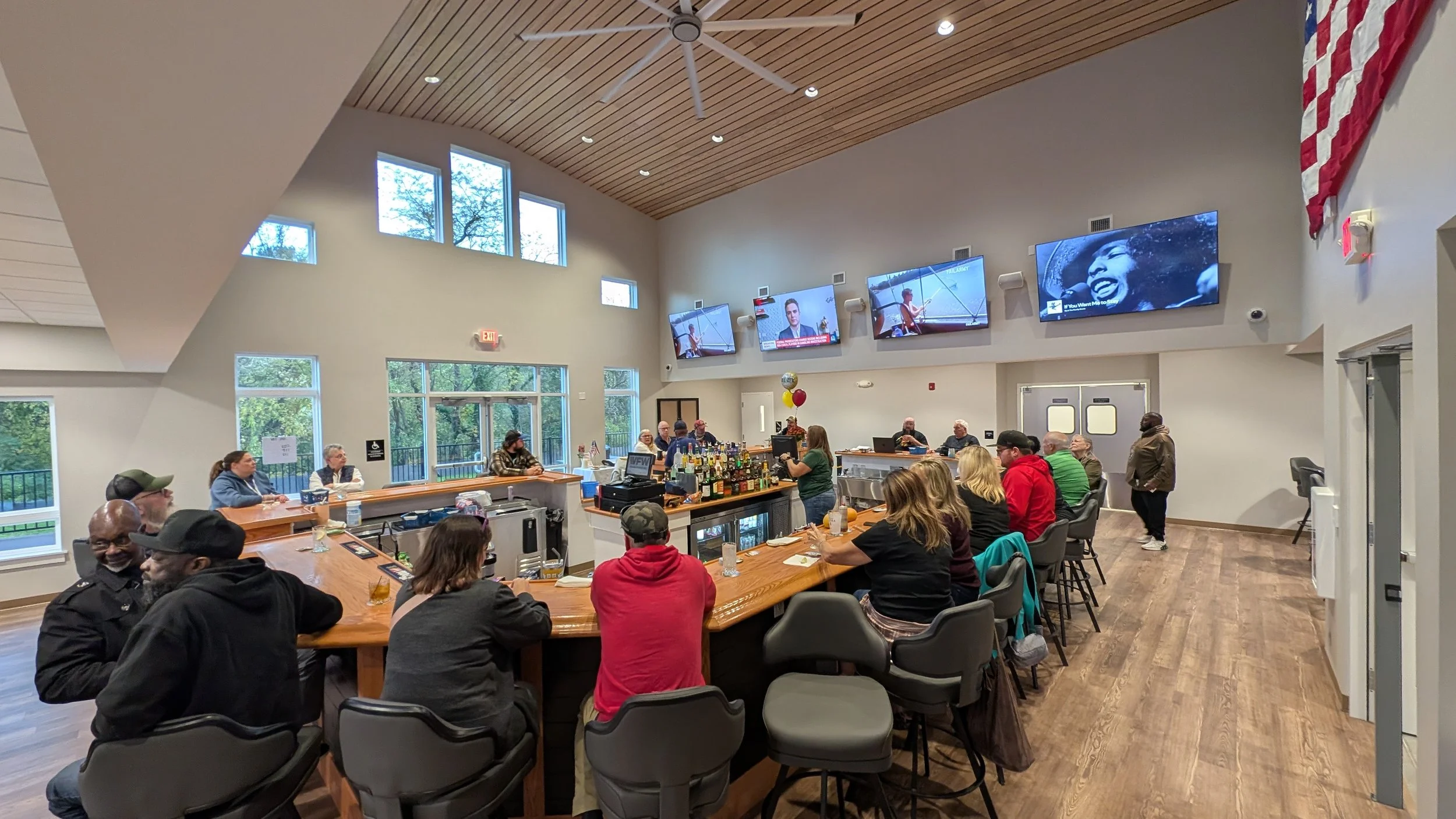 Interior of a bar or lounge with a curved wooden bar, multiple people sitting and standing, large windows, and several wall-mounted TVs playing different programs.