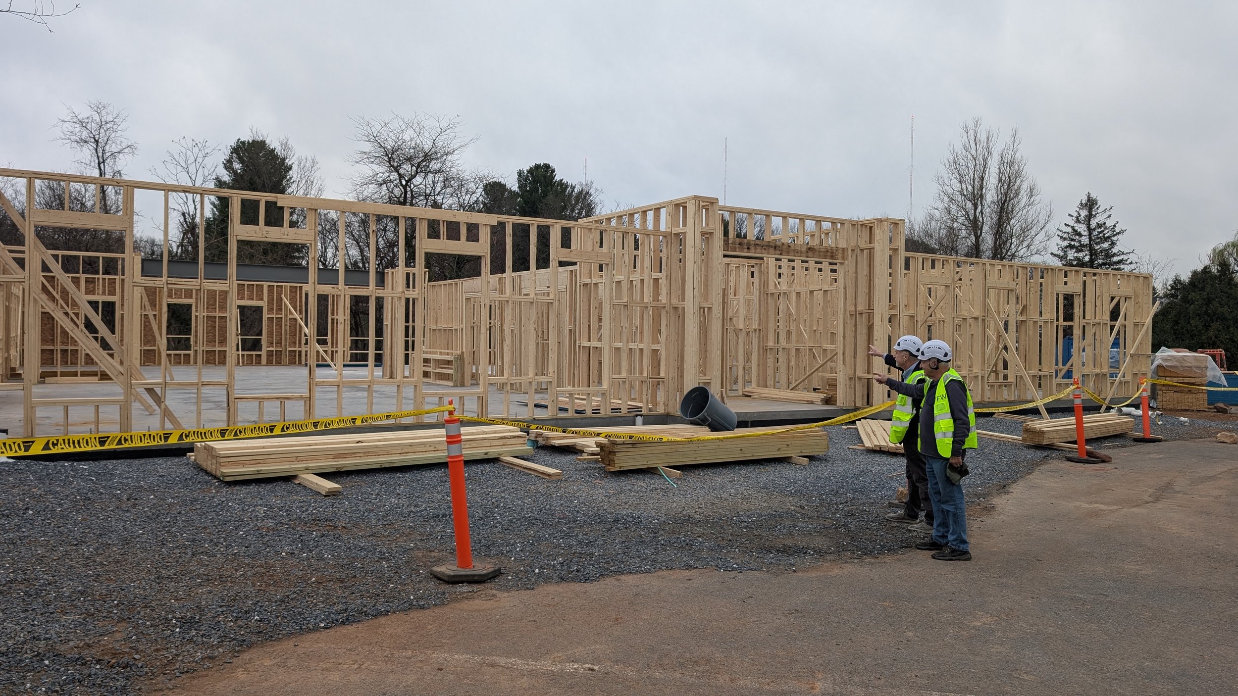 Construction site with wooden framing for building, three workers in safety vests and helmets observing and pointing, caution tape and building materials around.