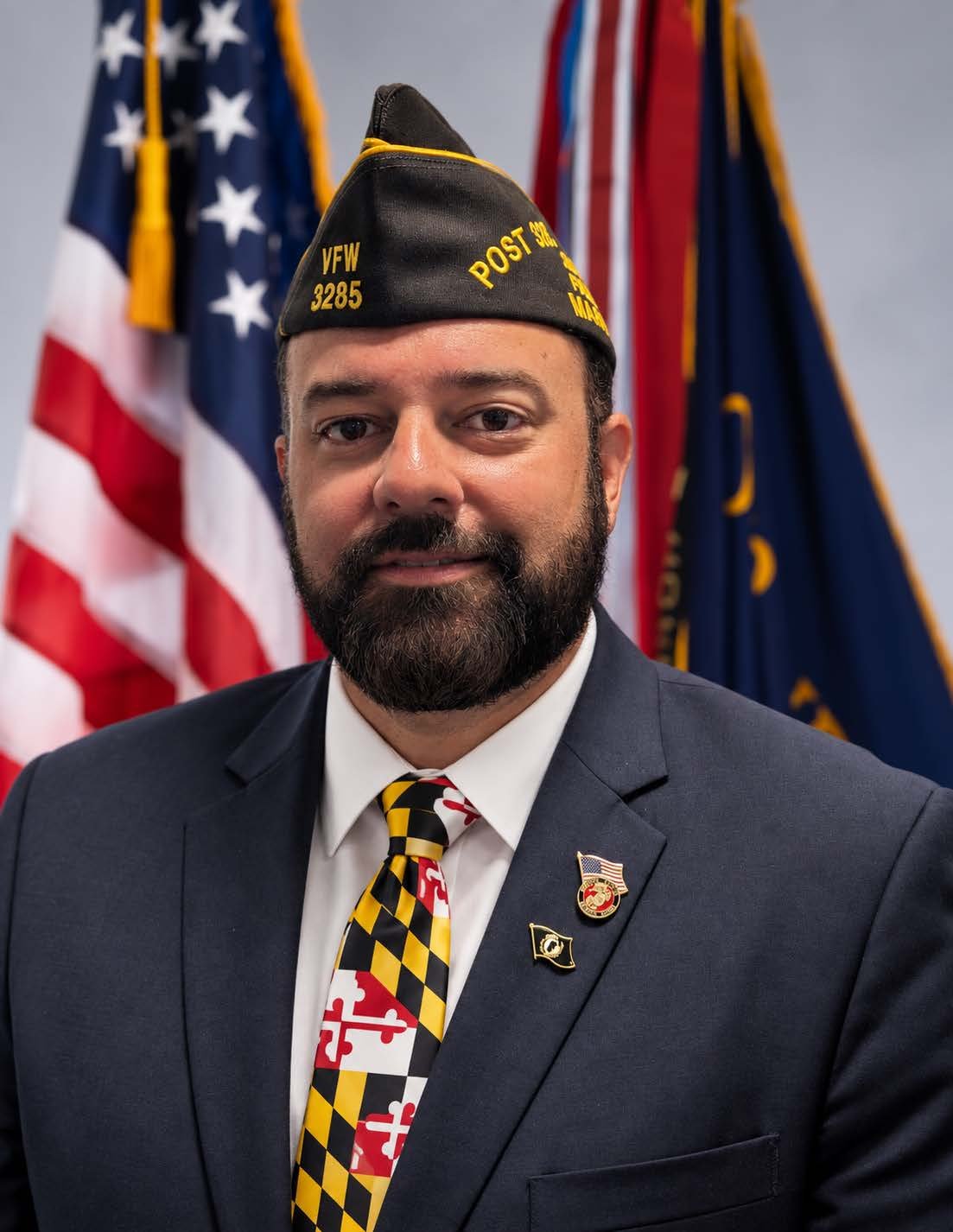 A man dressed in a suit with a U.S. flag pin and a veterans lapel pin, wearing a vintage veteran hat, standing in front of an American flag.