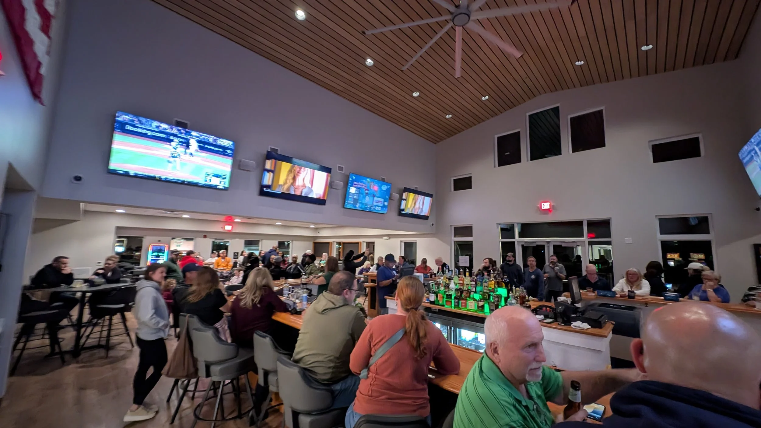 Inside a lively bar with patrons sitting at the bar or tables, watching multiple TVs on the wall showing sports and other programming, and socializing.