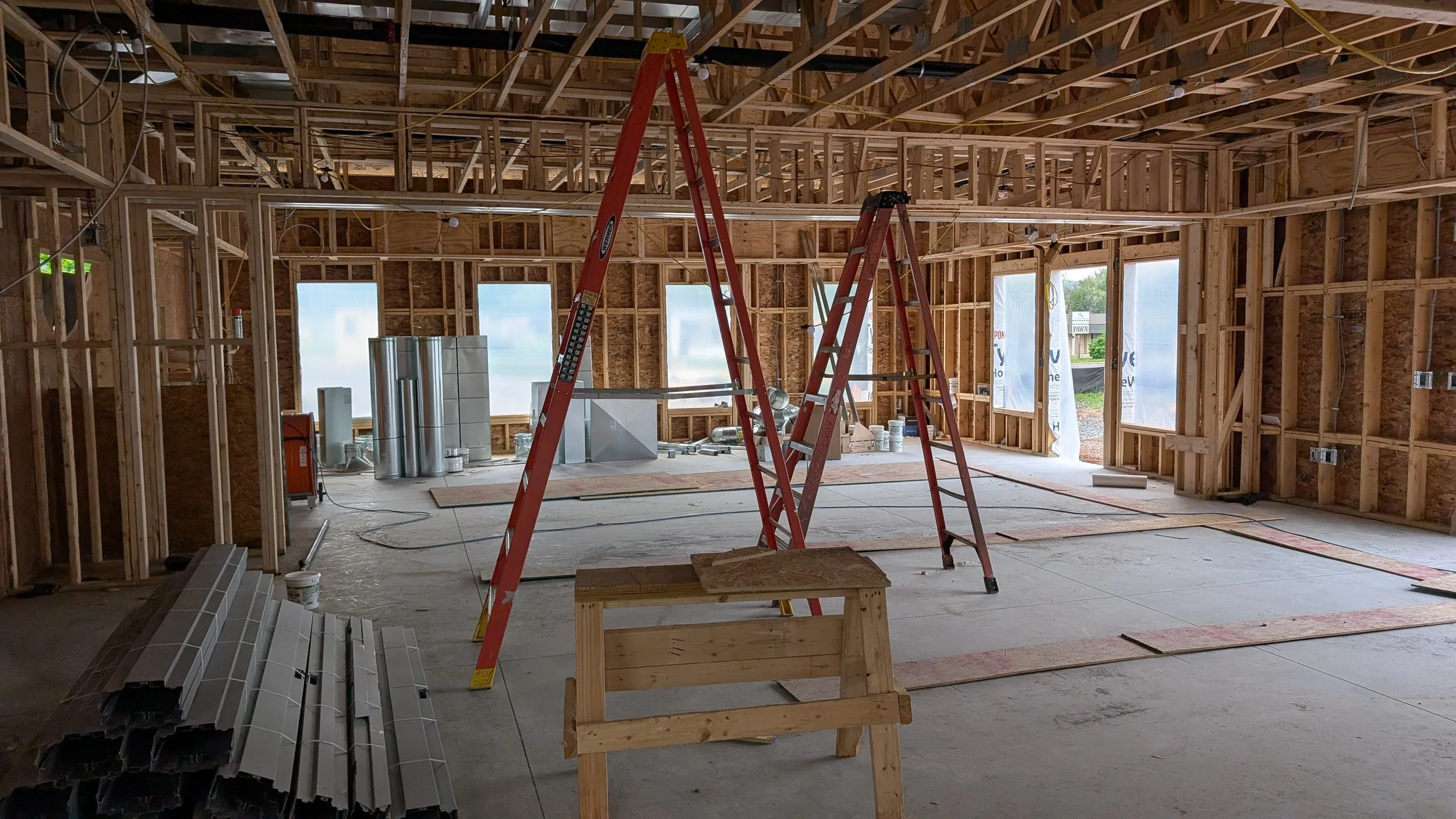 Interior view of a house under construction with exposed wooden framing, two red ladders, construction materials, and large windows.