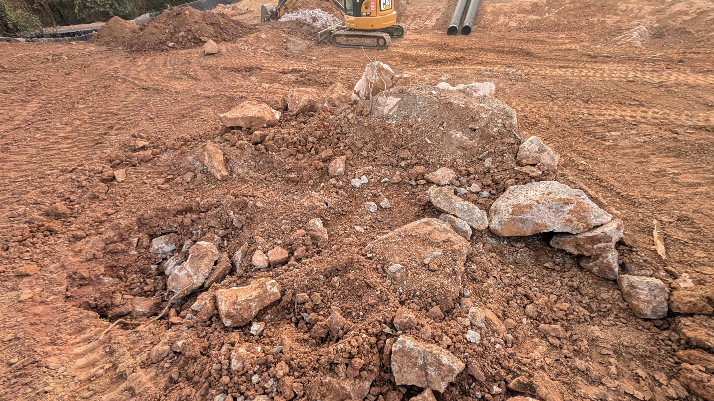 Construction site with dirt and rocks, a small yellow excavator in the background, and metal pipes near the top right corner.
