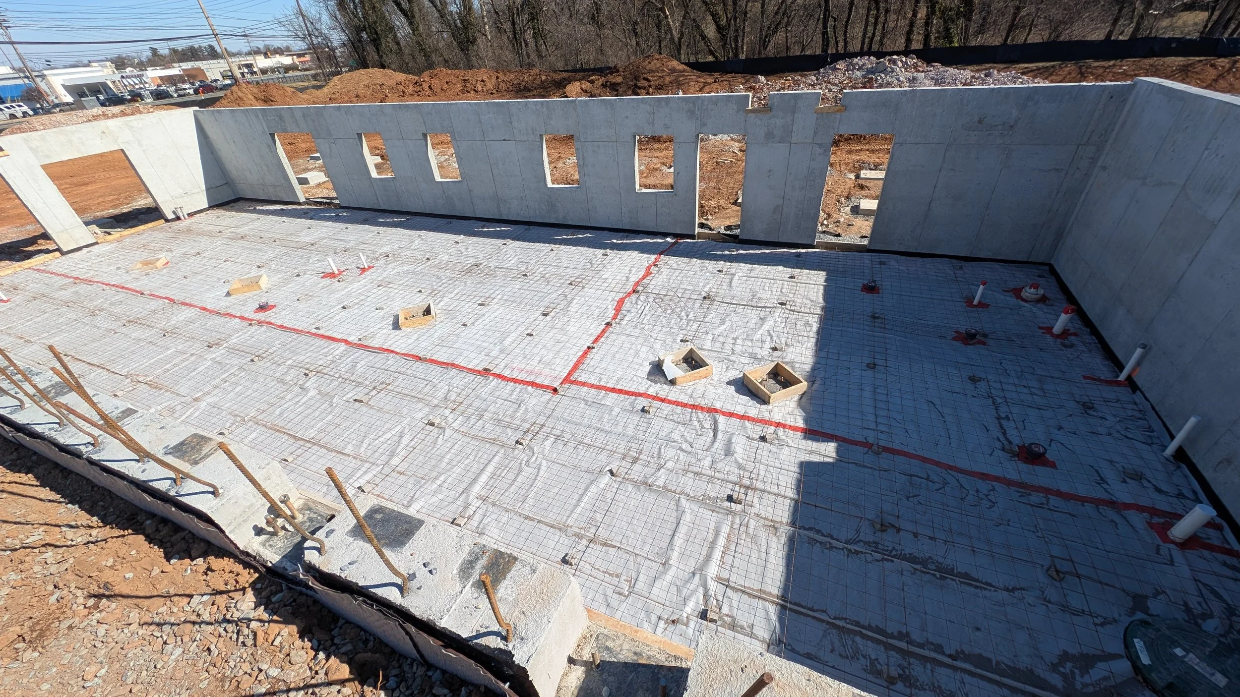 Construction site with a concrete foundation and walls, with rebar and plumbing pipes visible, during early building stages.