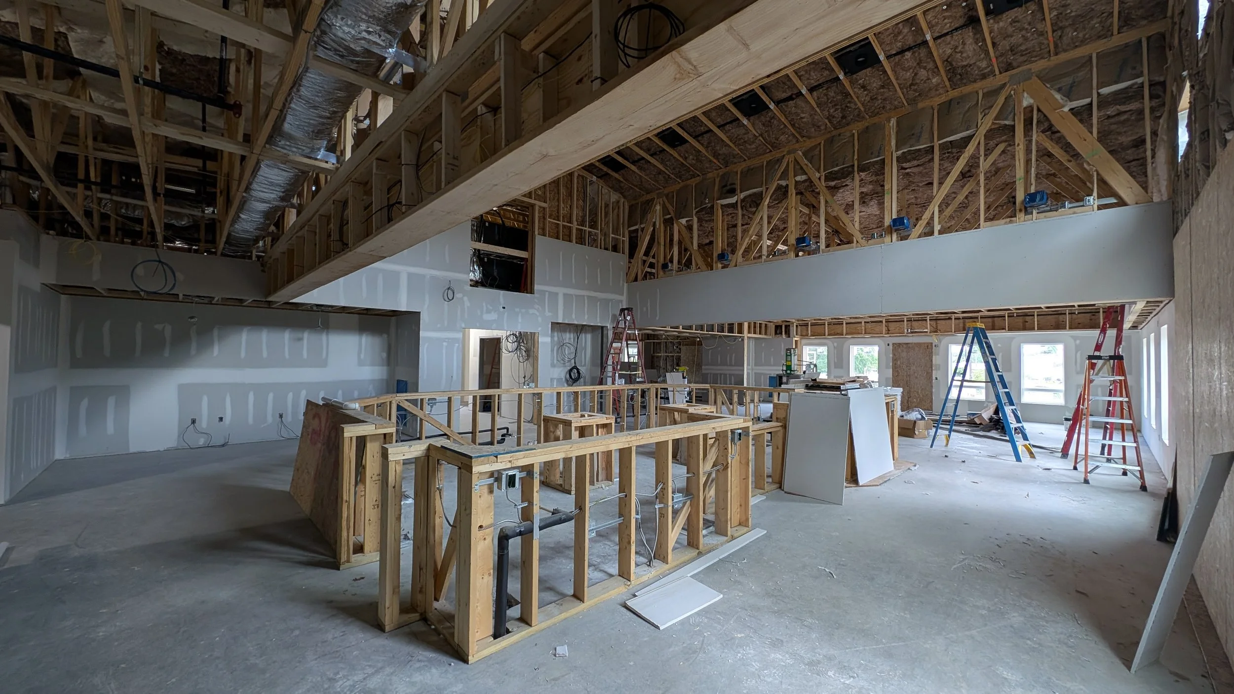 Interior of a building under construction with framed walls, exposed wooden beams, and ladders, some windows visible on the right side.
