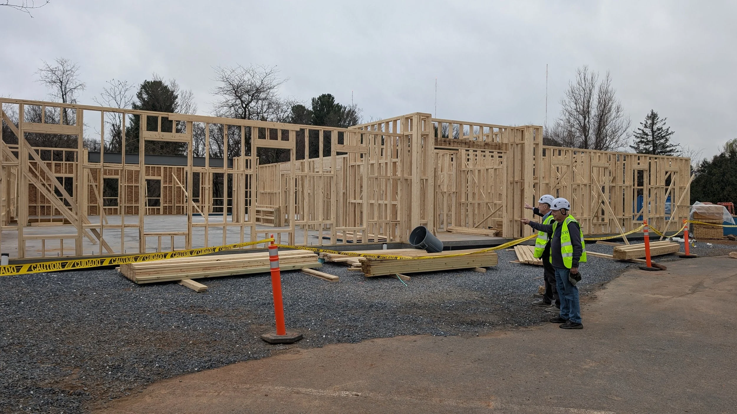 Three construction workers in safety vests and helmets standing at a construction site with wooden framing for a building. There are stacks of lumber, caution tape, and orange traffic cones in the foreground. Trees and a cloudy sky are in the backgro