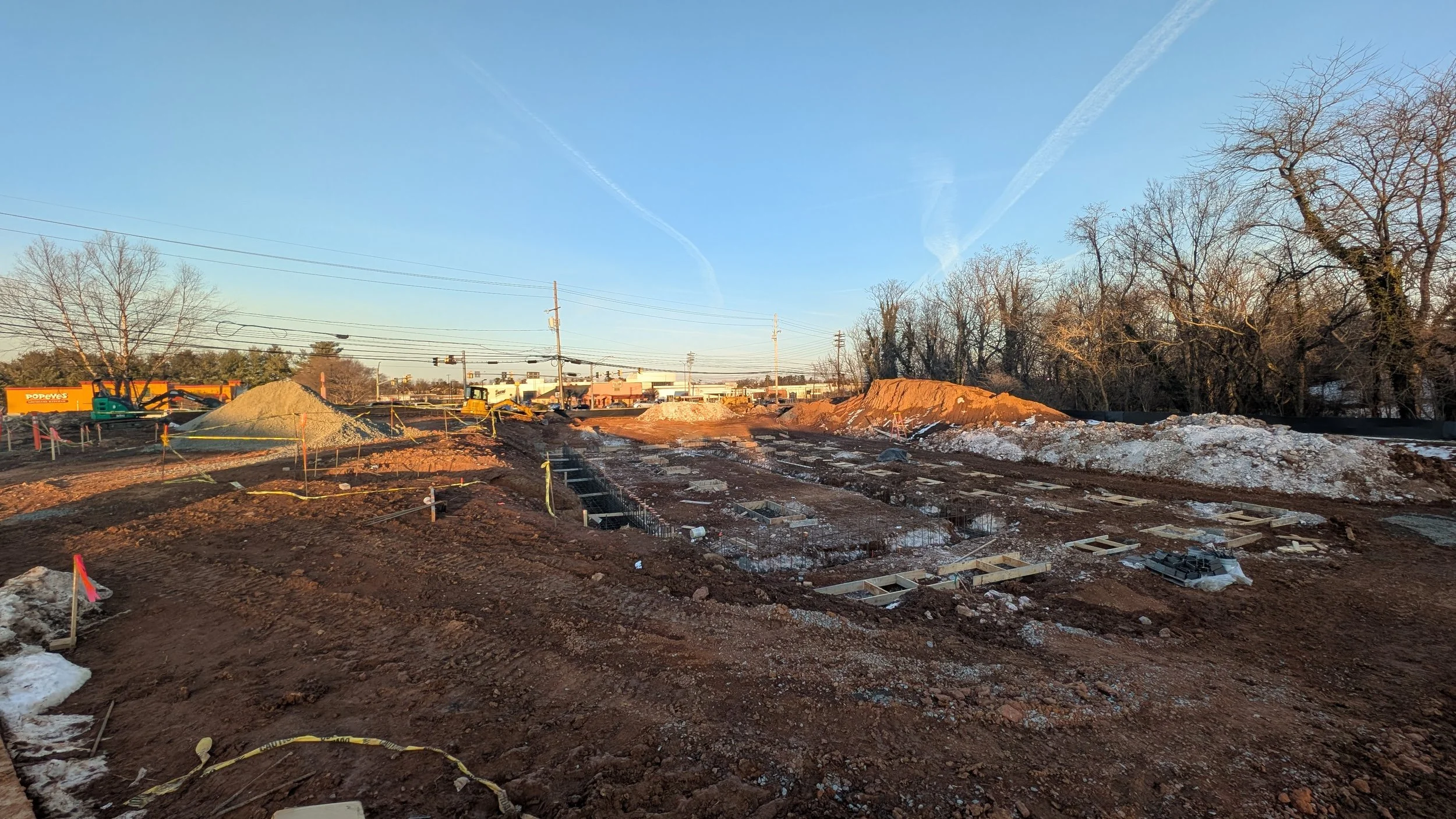 Construction site with dug-up earth, concrete foundation outlines, piles of dirt, construction equipment, and trees in the background under a clear sky.