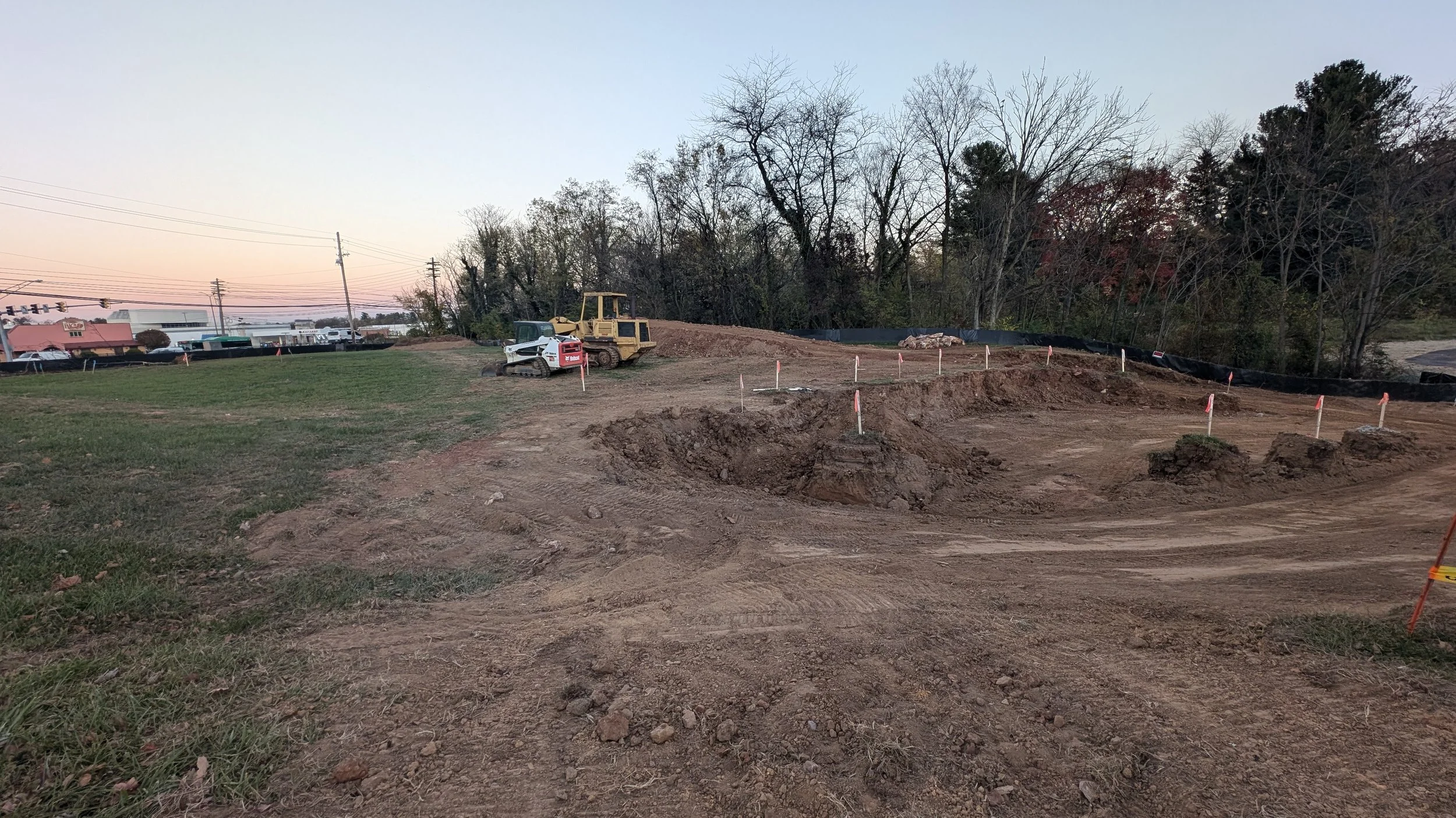 Construction site with dirt mounds, construction vehicle, and marked areas, alongside trees and power lines at dusk.