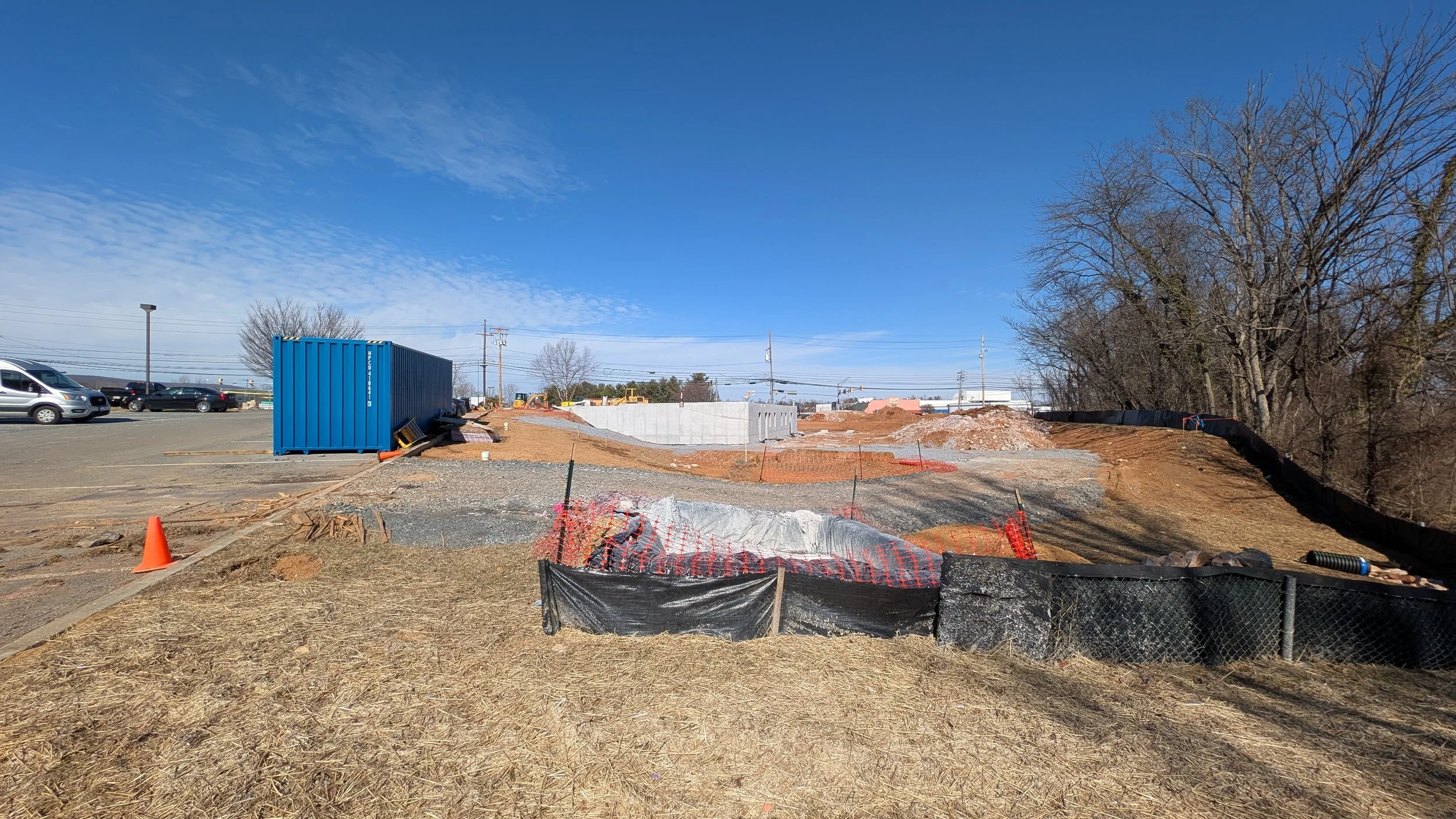 Construction site with orange safety fencing, construction equipment, dirt piles, and a blue storage container under a clear blue sky.