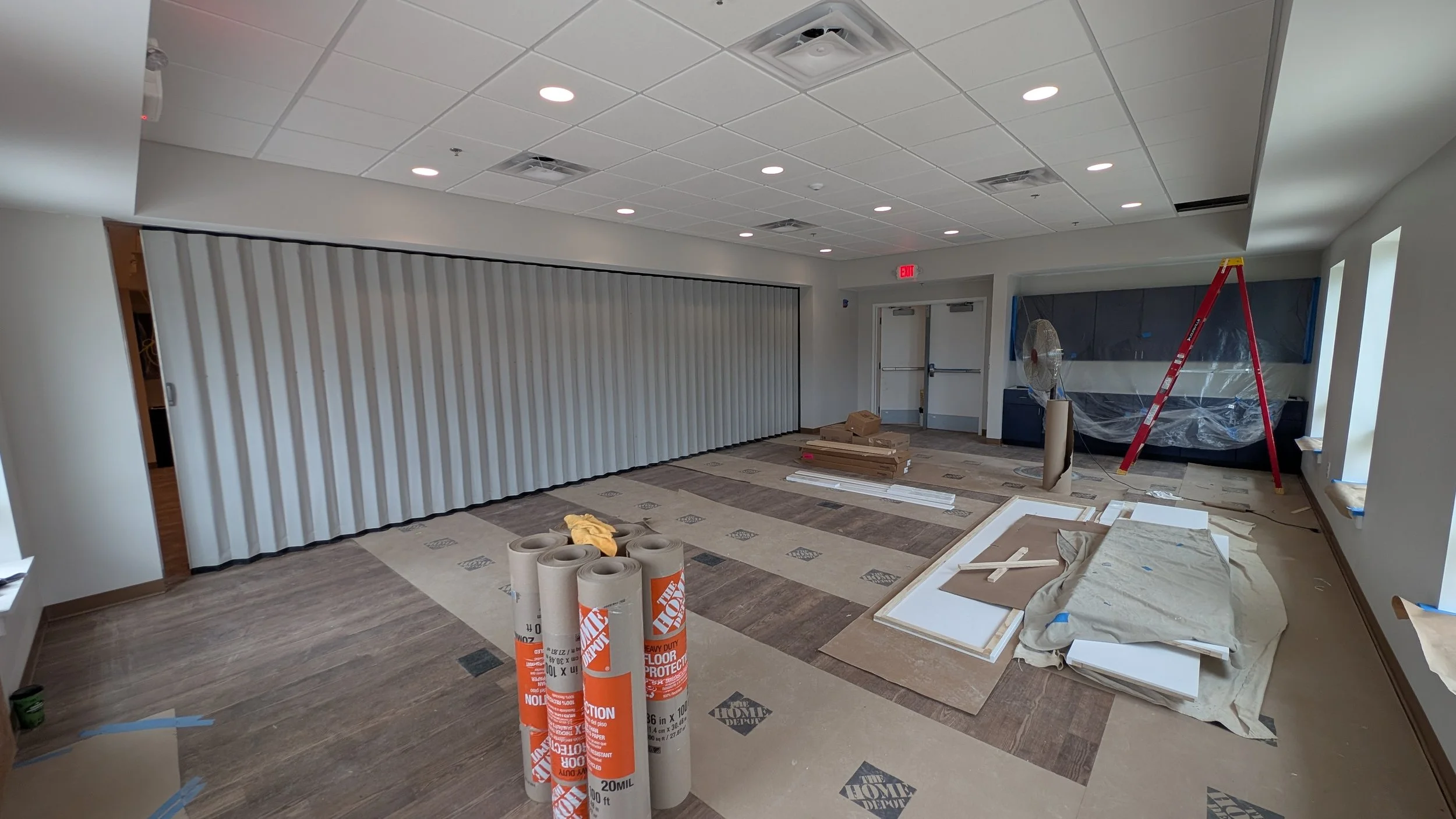 Interior room under construction with rolled flooring, ladder, fan, and construction materials, and a large wall covered with a privacy curtain.