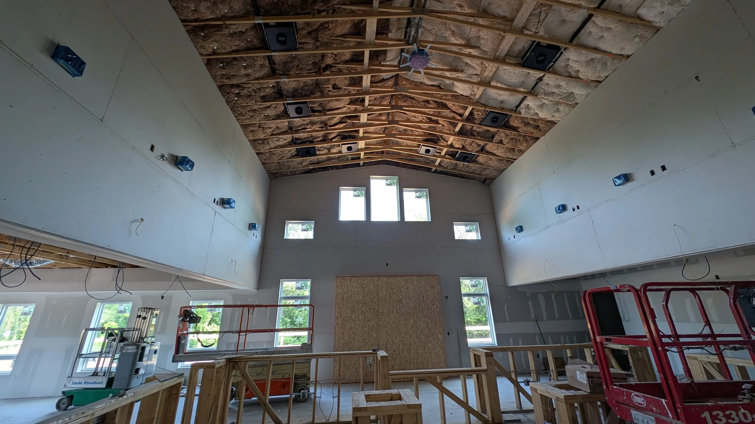 Interior of a building under construction, showing a high ceiling with exposed insulation and wooden beams, multiple windows on the front wall, and construction equipment and scaffolding inside.