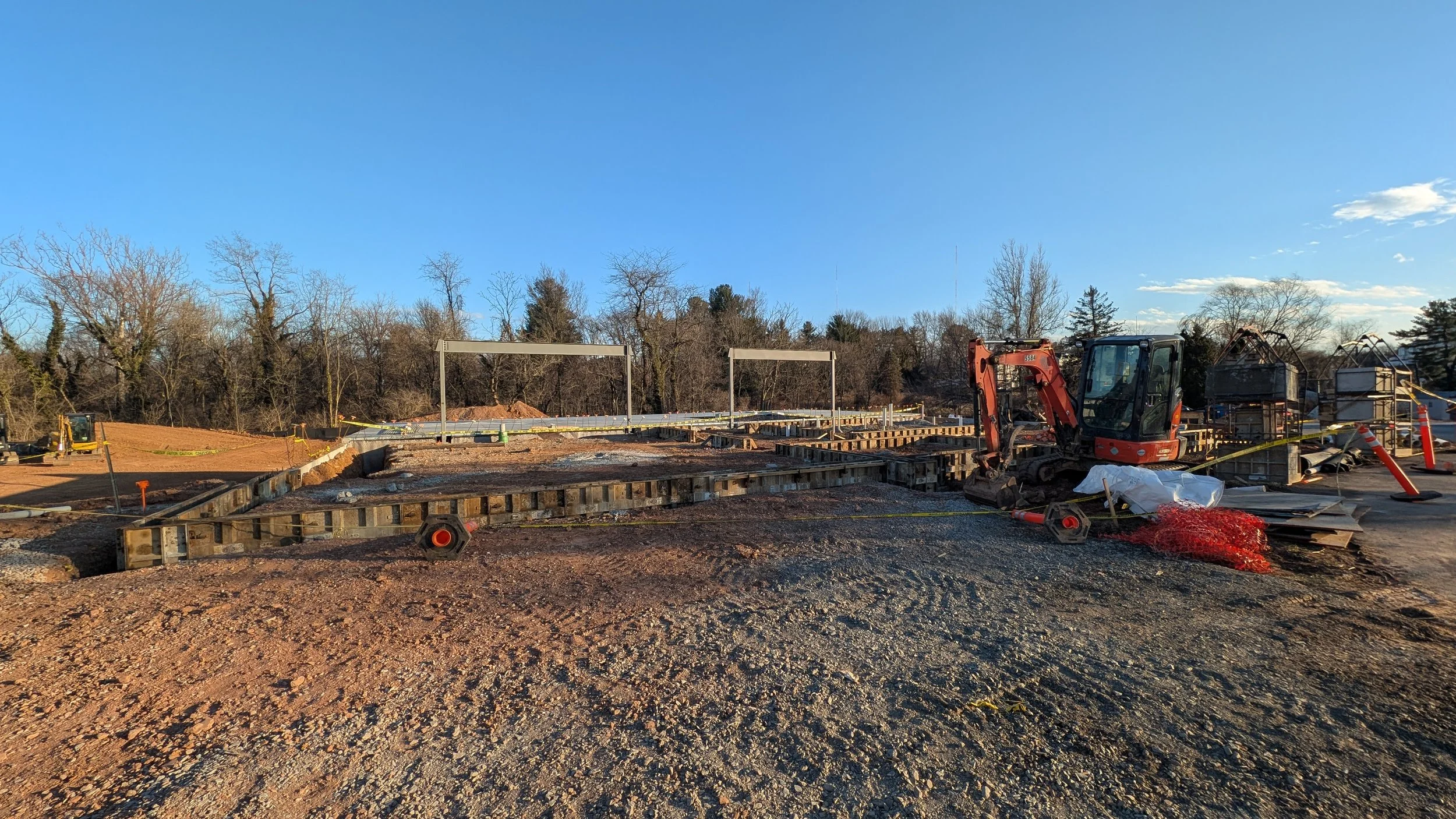 Construction site with a small excavator, framing for a foundation, orange safety barriers, and construction equipment under a blue sky.