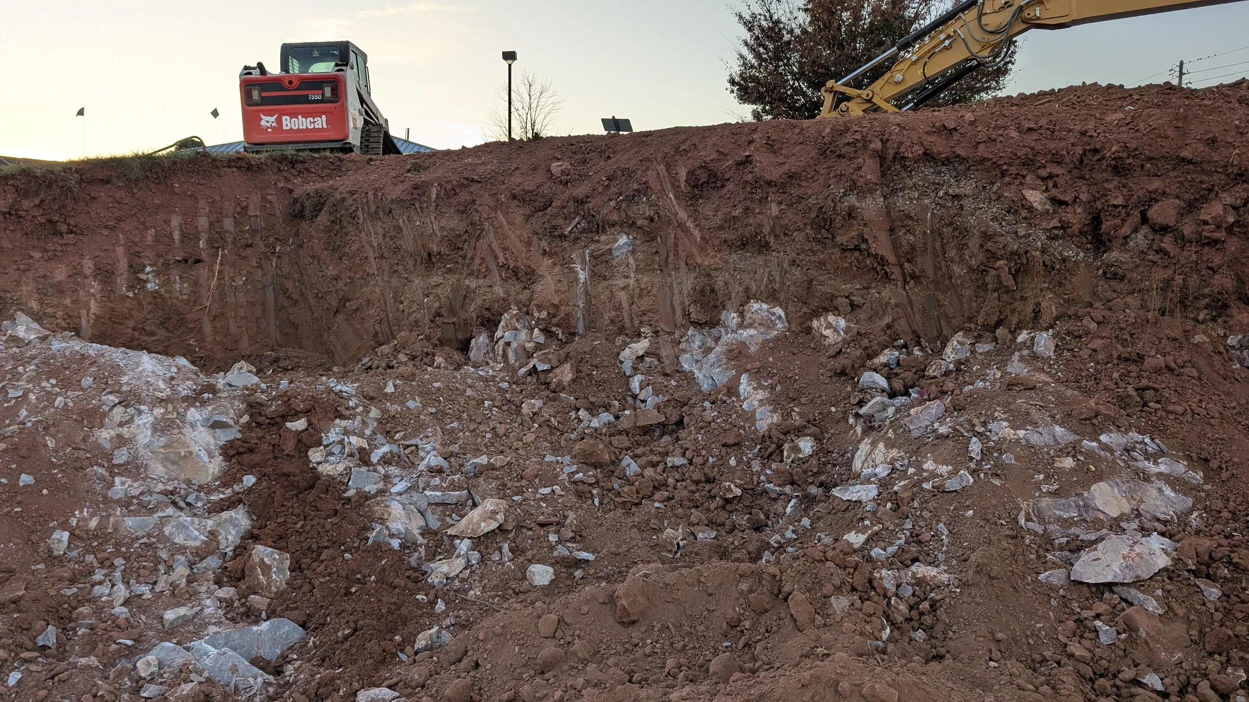 Construction site with an excavator digging a deep trench. The construction equipment, labeled 'Bobcat,' is visible on top of the slope, and part of the excavator arm can be seen on the right side of the image.