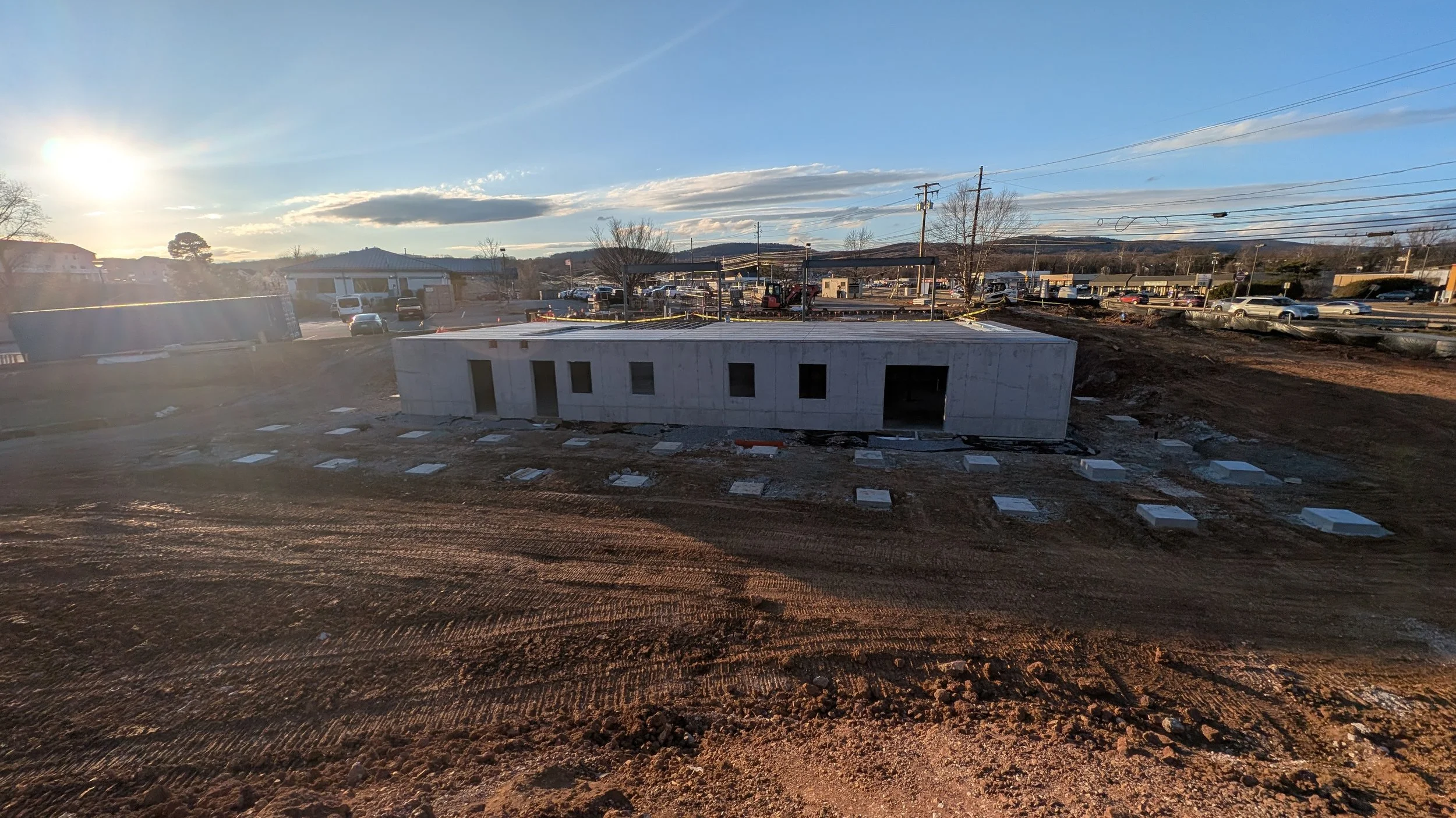 Construction site with a partially built concrete structure, surrounded by dirt and construction materials, under a clear sky with the sun setting in the background.