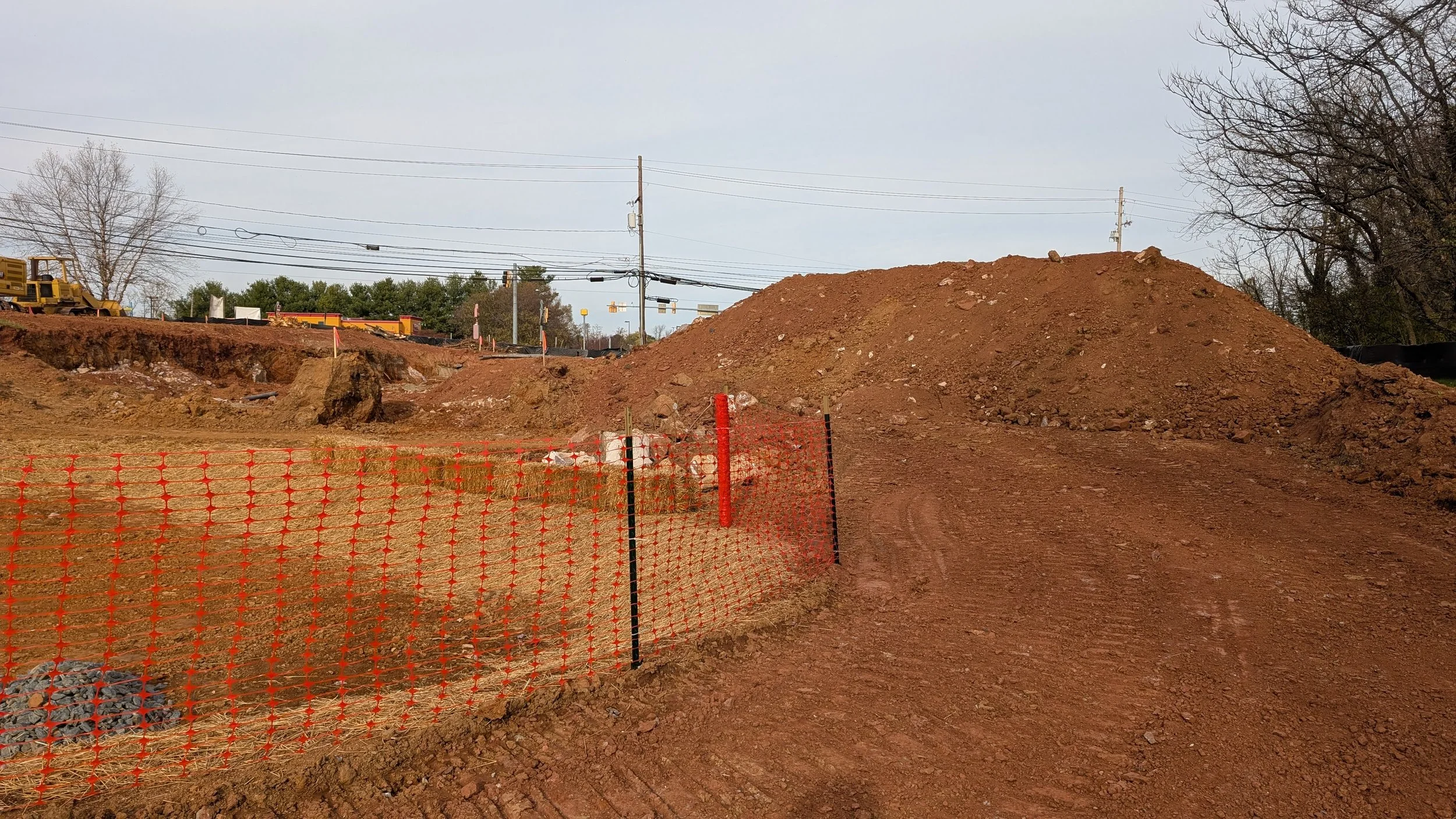 Construction site with a dirt pathway, orange safety fence, piles of soil, and construction equipment in the background under an overcast sky.